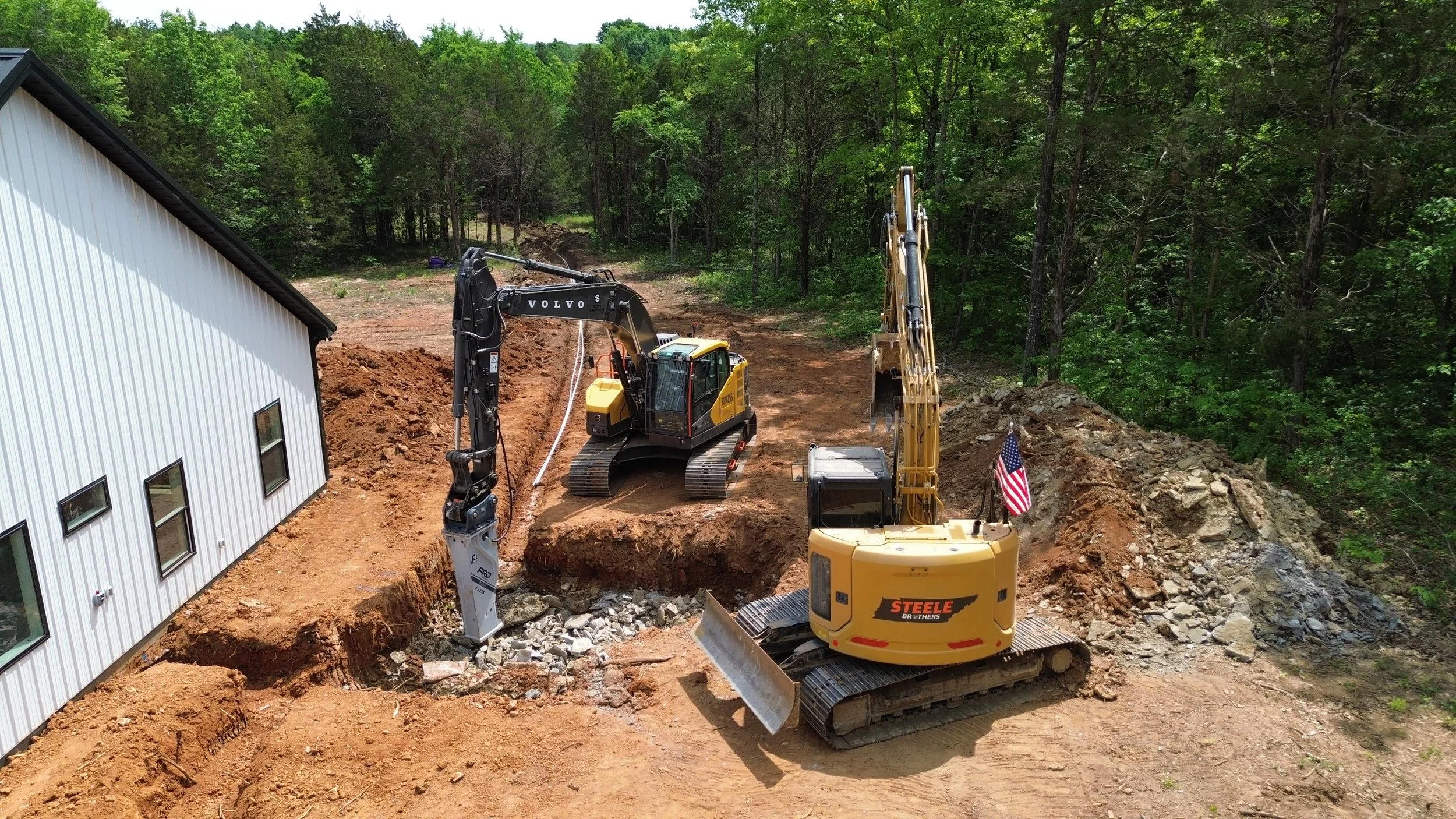 Construction site with excavators digging and moving earth near a white building with trees in the background.