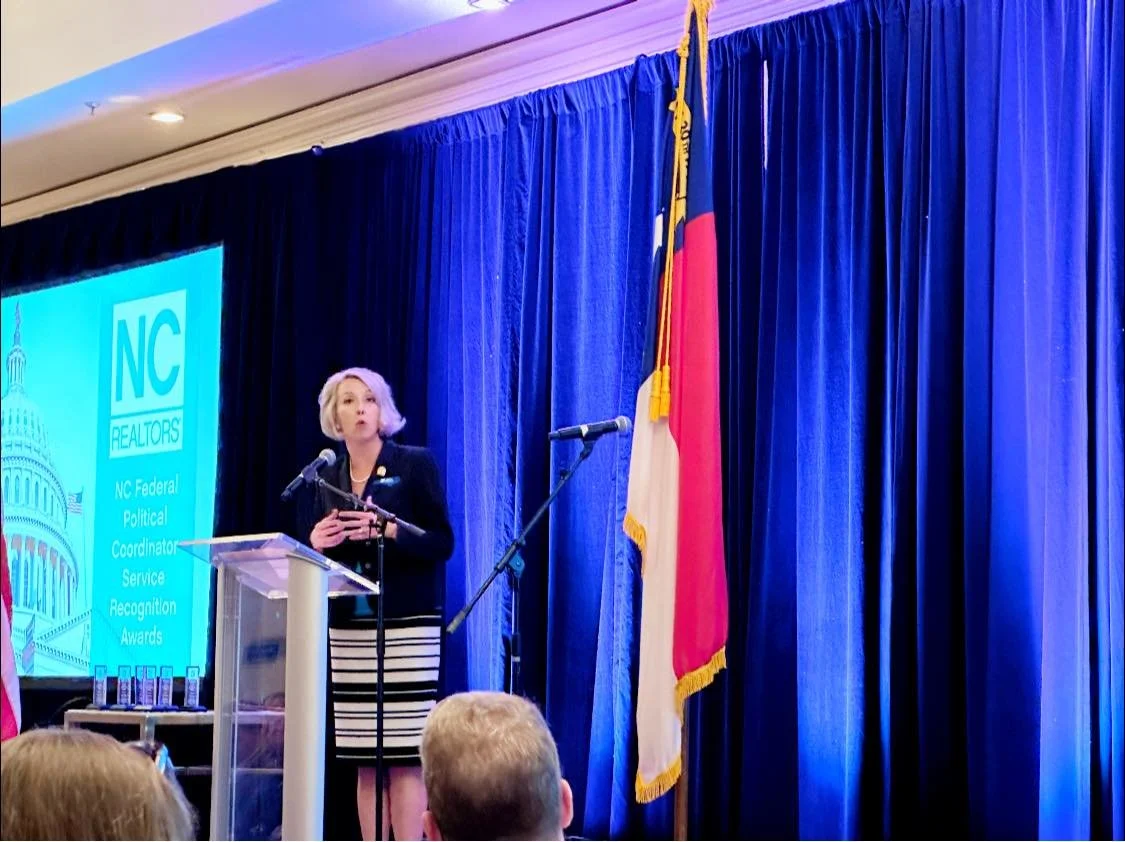 A woman with blonde hair in a purple blazer and striped skirt speaks at a podium during an awards ceremony. Behind her, a large screen displays the NC Realtors logo and a headline about a political recognition award. The stage has blue curtains and an American flag. Audience members are visible in foreground.