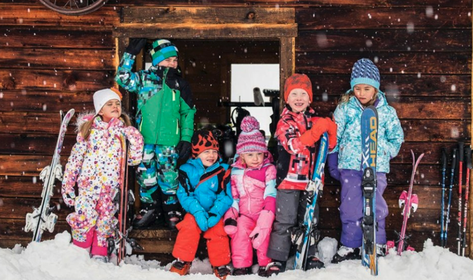 Group of children in winter clothing with skis and snowshoes, standing in front of a wooden cabin in snowy weather.