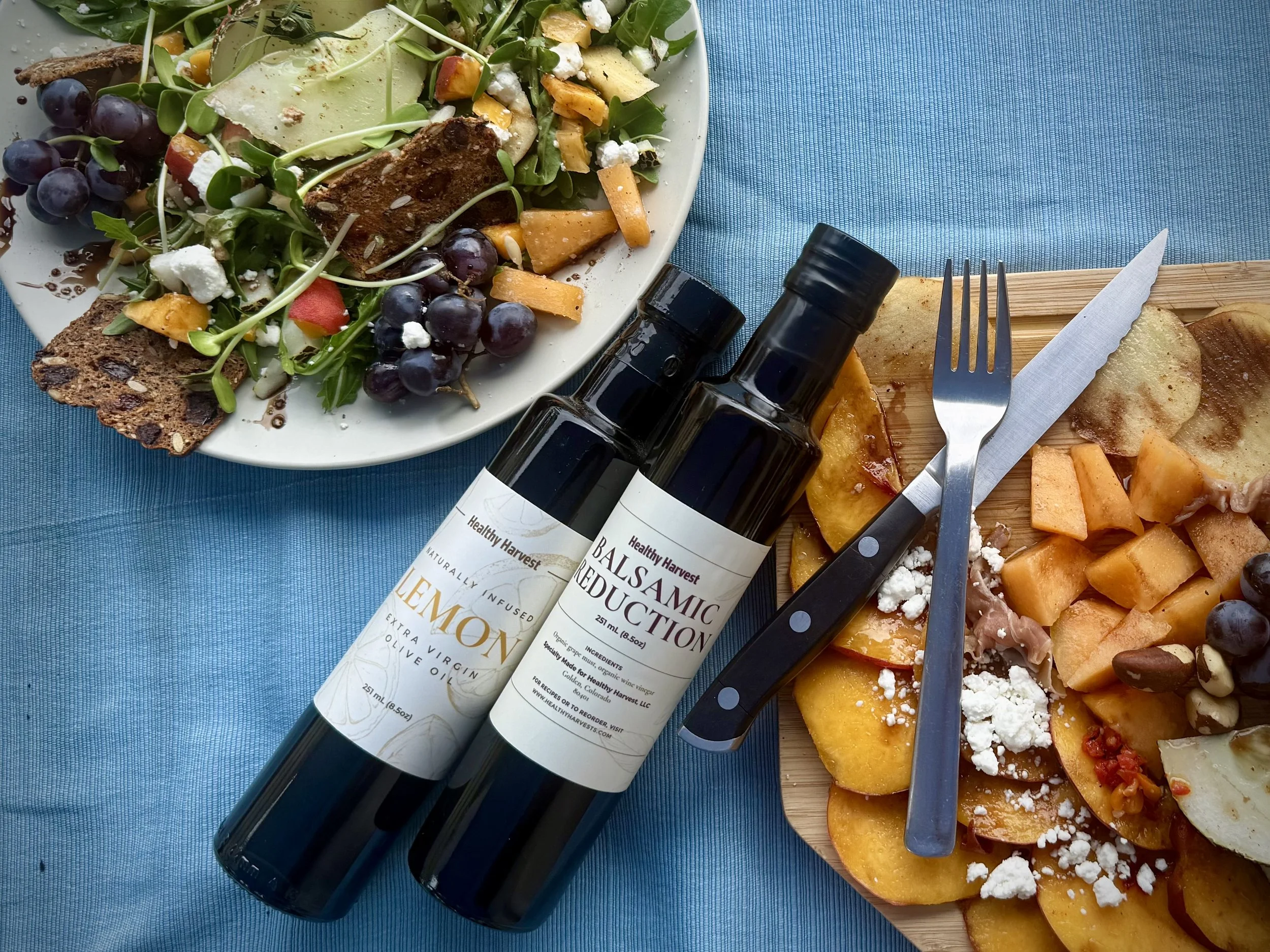 Aplate with roasted vegetables, cheese, and crackers, a salad with grapes, greens, and cheese, two bottles of balsamic reduction and lemon olive oil, a fork, knife, and napkin on a wooden cutting board, all on a blue tablecloth.