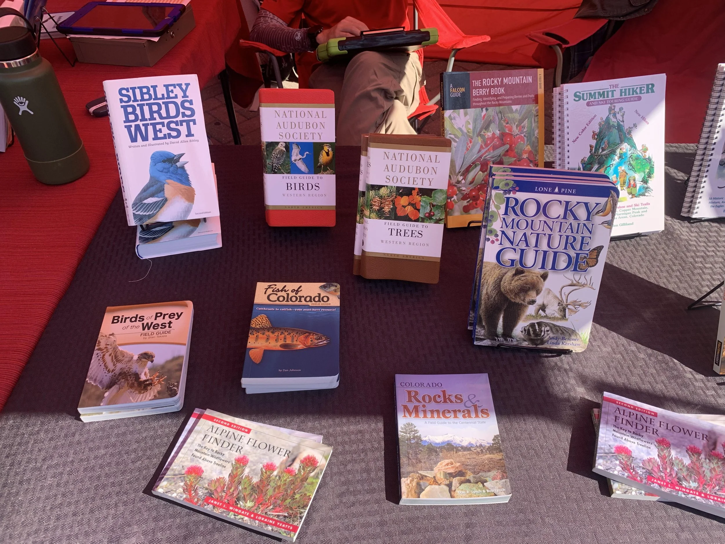A table displaying various nature and wildlife books, including guides on birds, trees, rocks, minerals, and flowers, with a person sitting behind using a tablet.