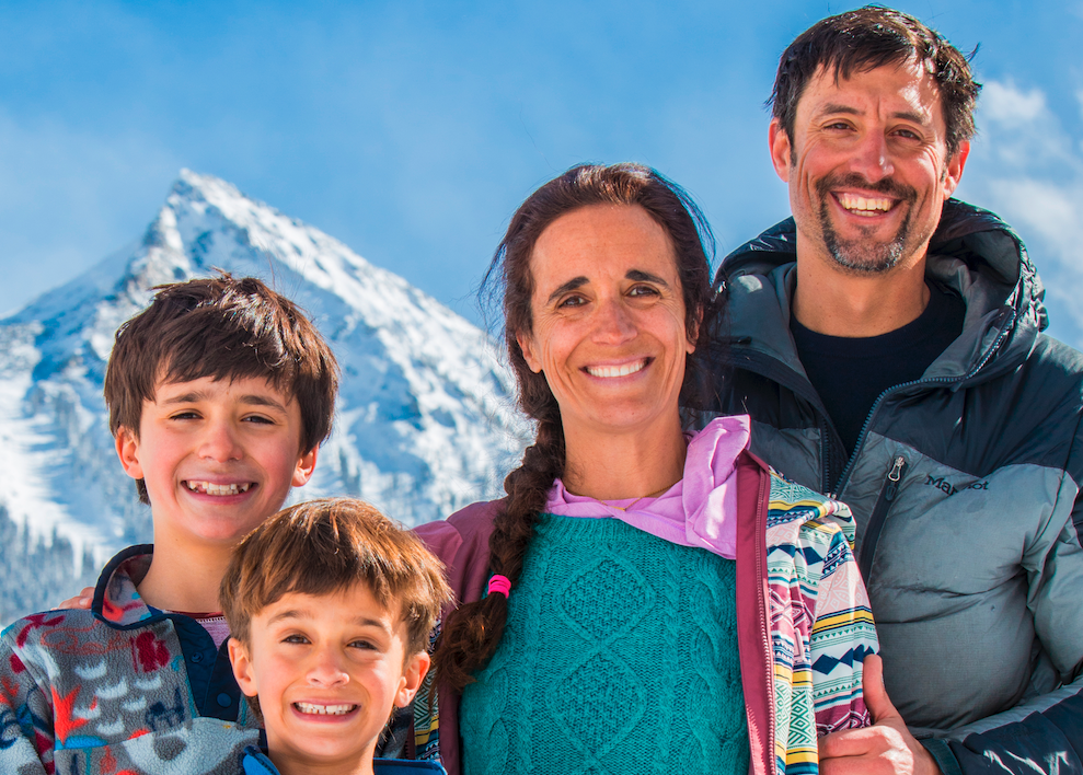 Family of four smiling outdoors with snow-covered mountains in the background.