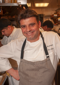 Chef smiling in a busy kitchen wearing a white chef's coat and gray apron.