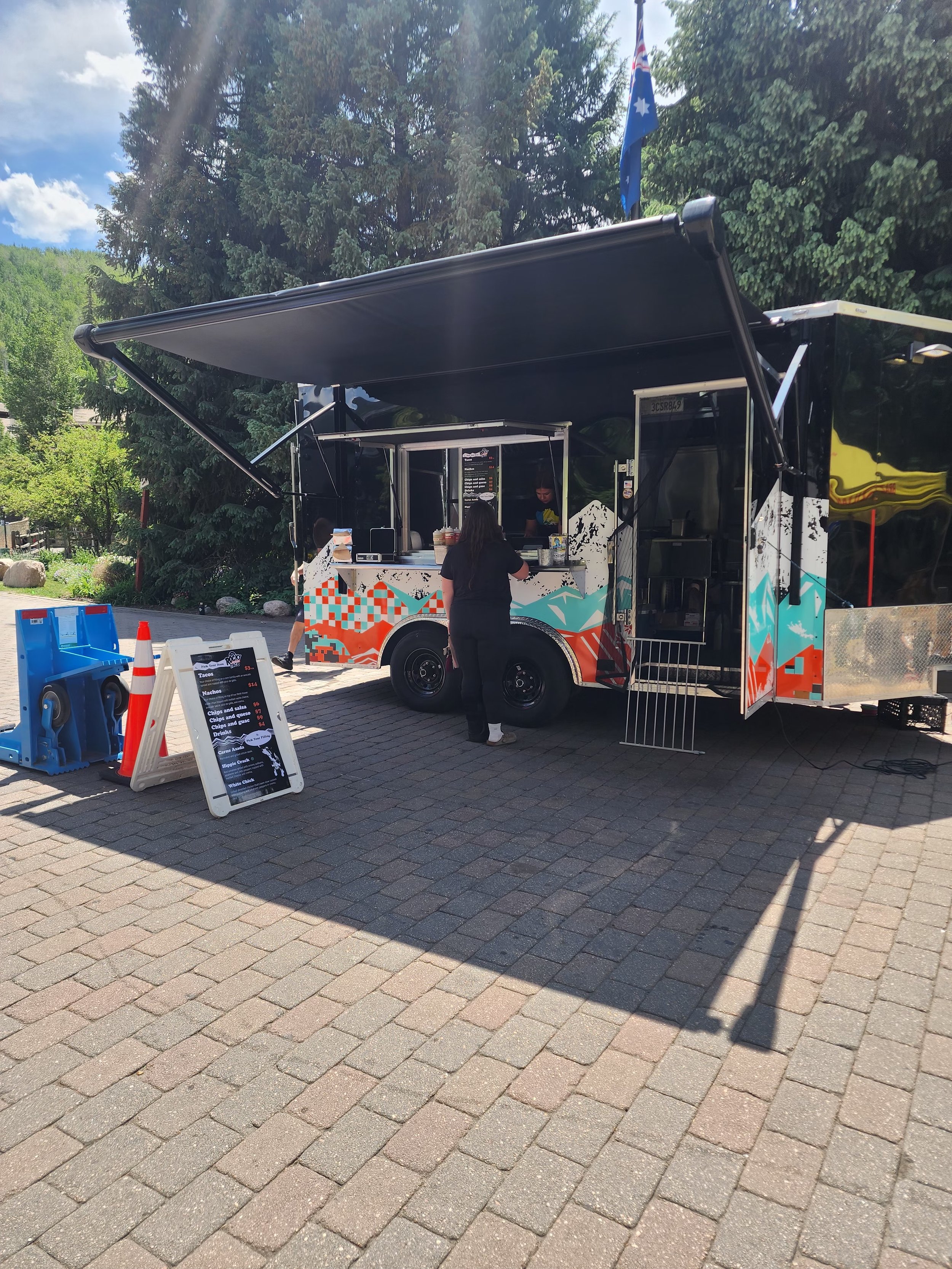 A colorful food truck serving tacos, with a woman ordering and a man preparing food inside; the truck is parked on a paved area with trees and a blue sky in the background.