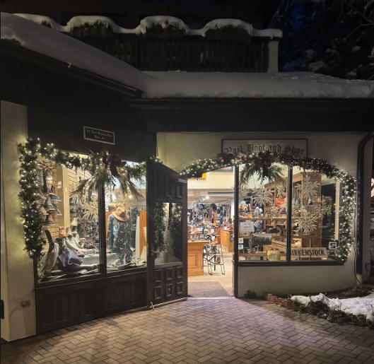 View of a store decorated with Christmas garlands and lights, with snow on the roof and outside, open for browsing.