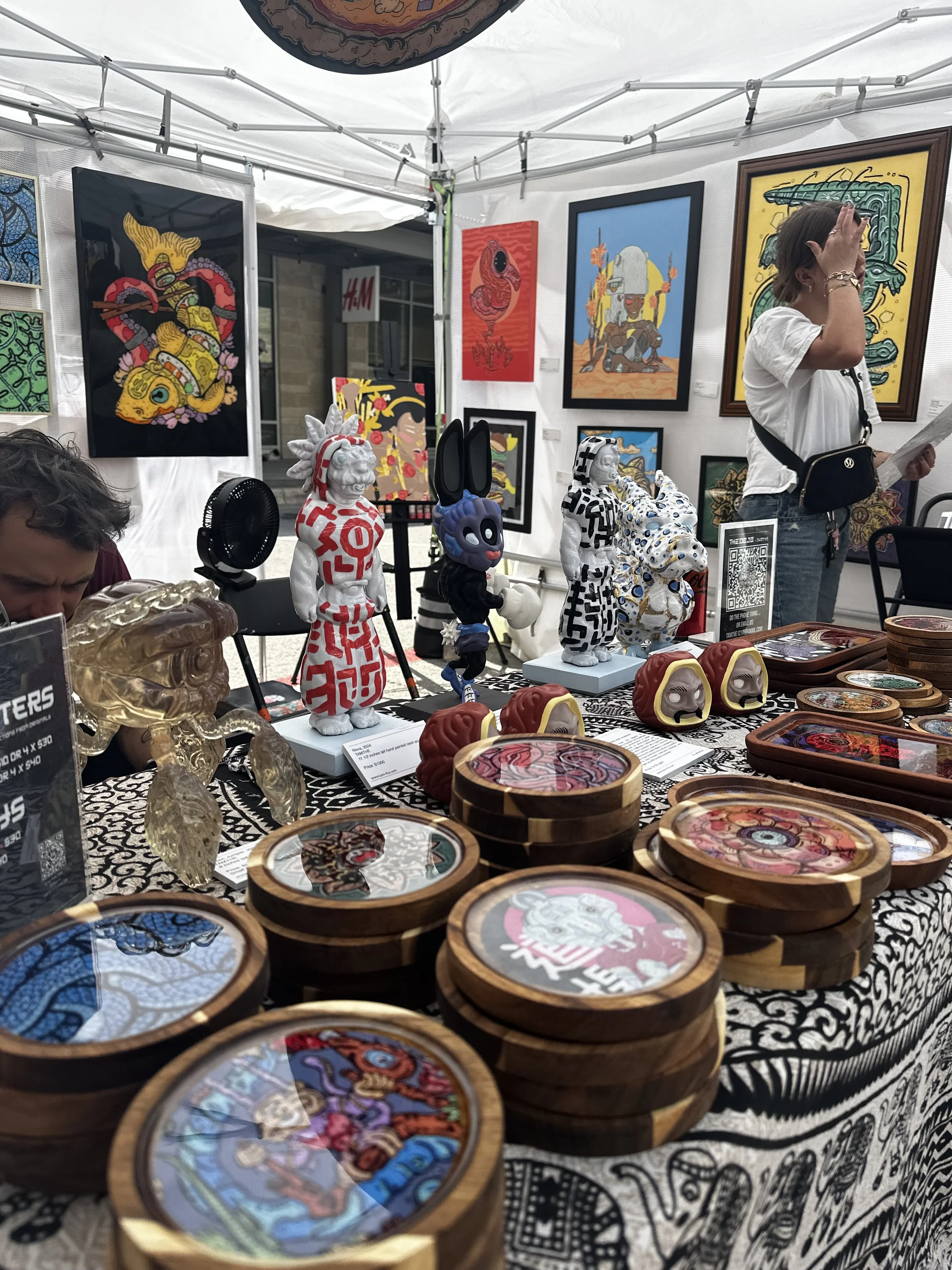 Art booth at an outdoor market displaying colorful artwork and sculptures, with people browsing and a woman reading a paper.