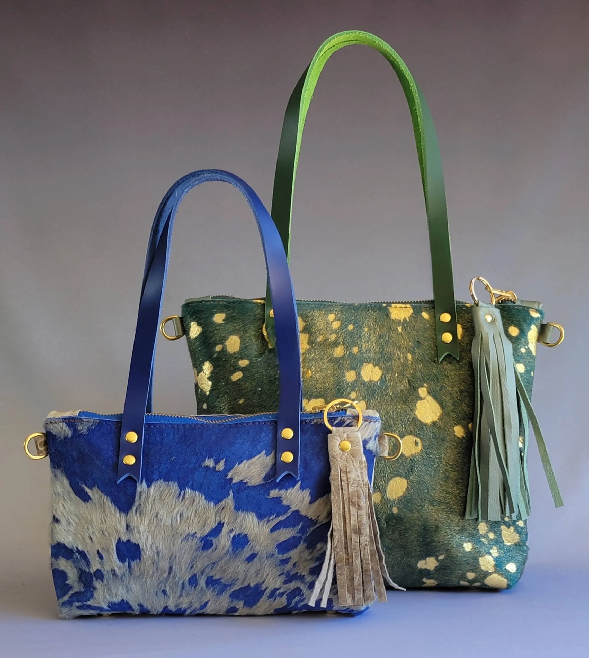 Two handbags with colorful handles and spots, one blue with animal print and the other green with gold splatters, featuring decorative tassels and gold rings.