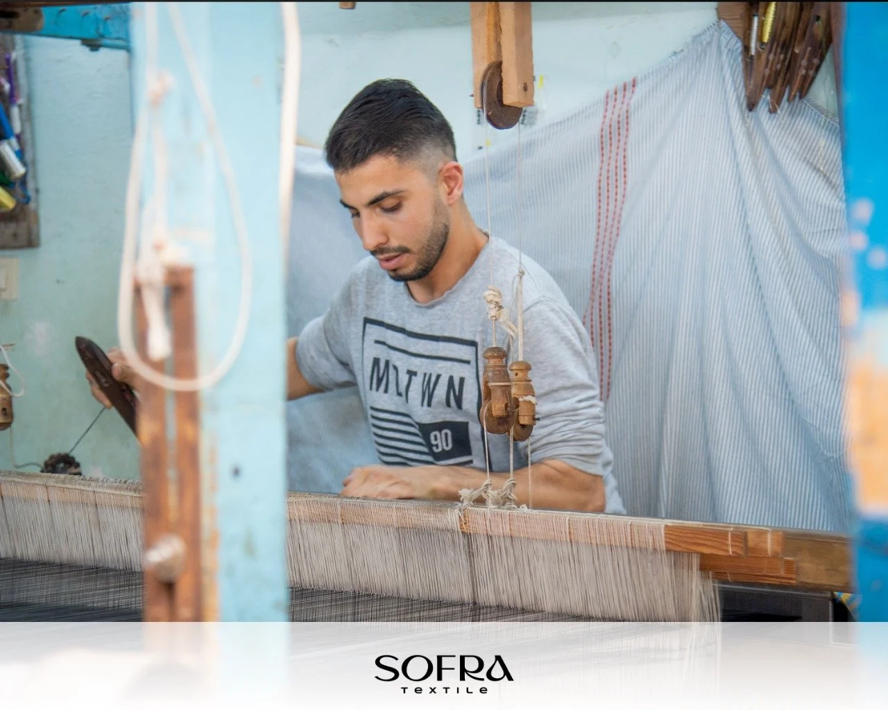 A young man working on a traditional textile loom in a workshop, wearing a light gray sweatshirt with black lettering.