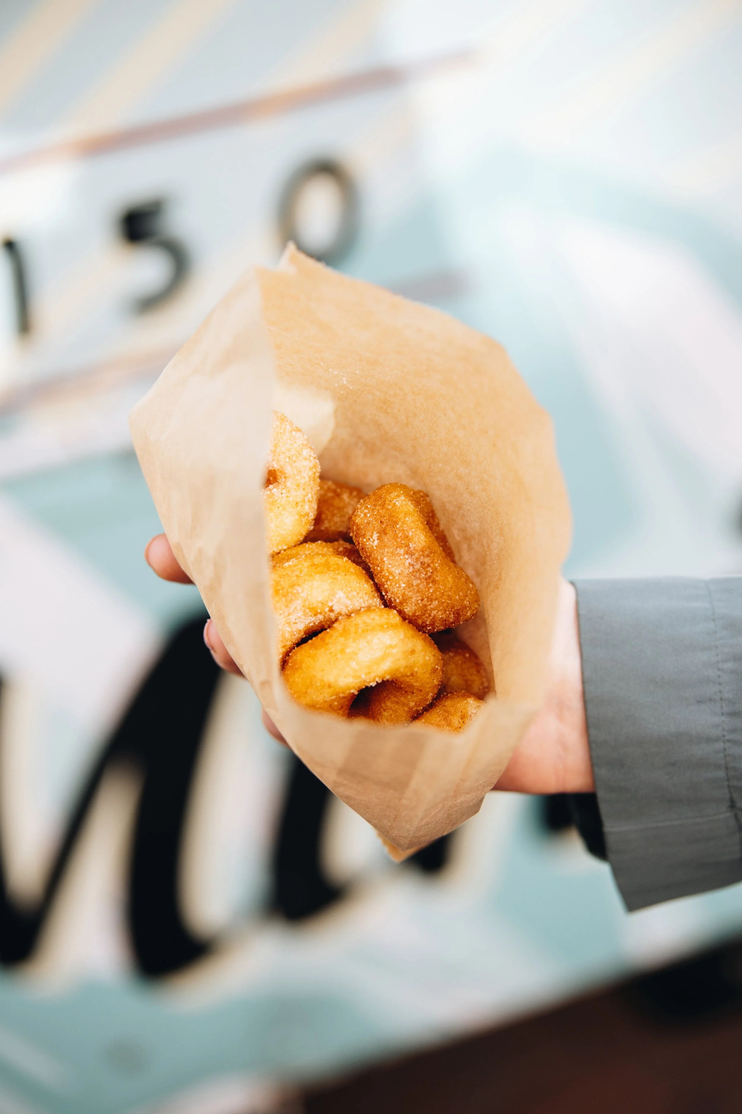 Hand holding a paper bag of cinnamon sugar coated churros in front of a bakery display counter.