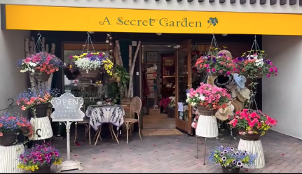 storefront of 'A Secret Garden' featuring hanging flower baskets, potted flowers, outdoor chairs, and tables with an 'Open' sign in the window