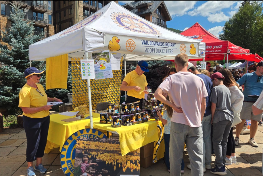 People gathering at a booth during an outdoor event, with a white canopy tent displaying a sign that reads 'Vail Edwards Rotary Adopt Ducks Here.' The booth has yellow decorations and rubber duck merchandise, with a person in a yellow shirt and blue hat selling ducks. Other tents and trees are visible in the background.