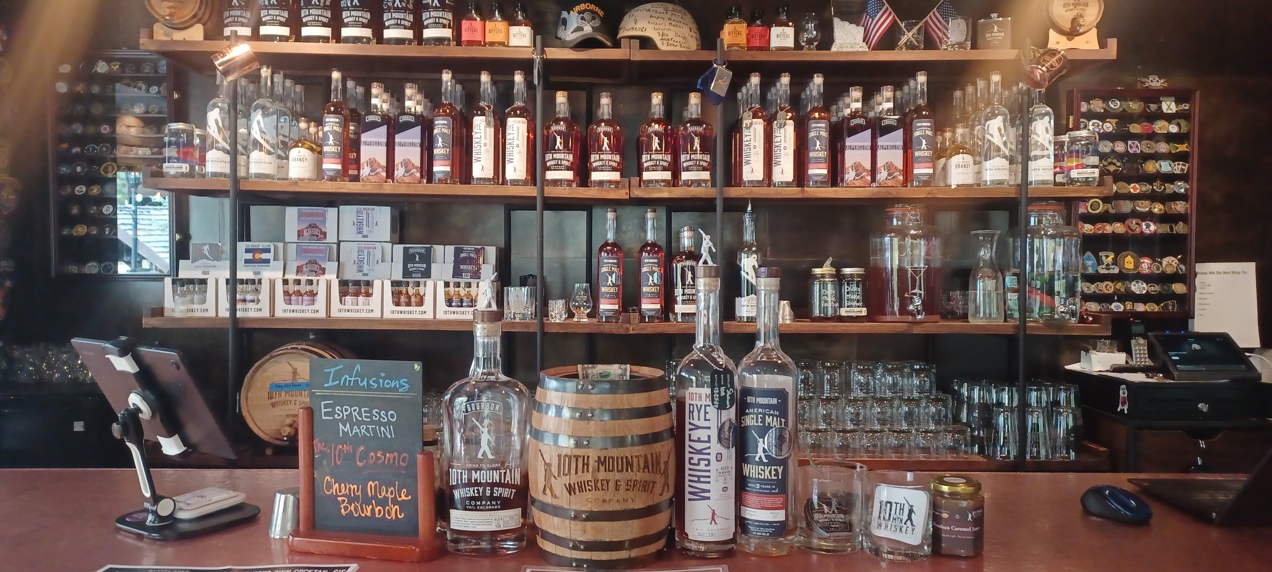 Bar counter with bottles of whiskey, glasses, and a chalkboard sign listing drinks like espresso martini, 10th Cosmo, and cherry maple bourbon, with shelves of additional liquor bottles and glassware in the background.