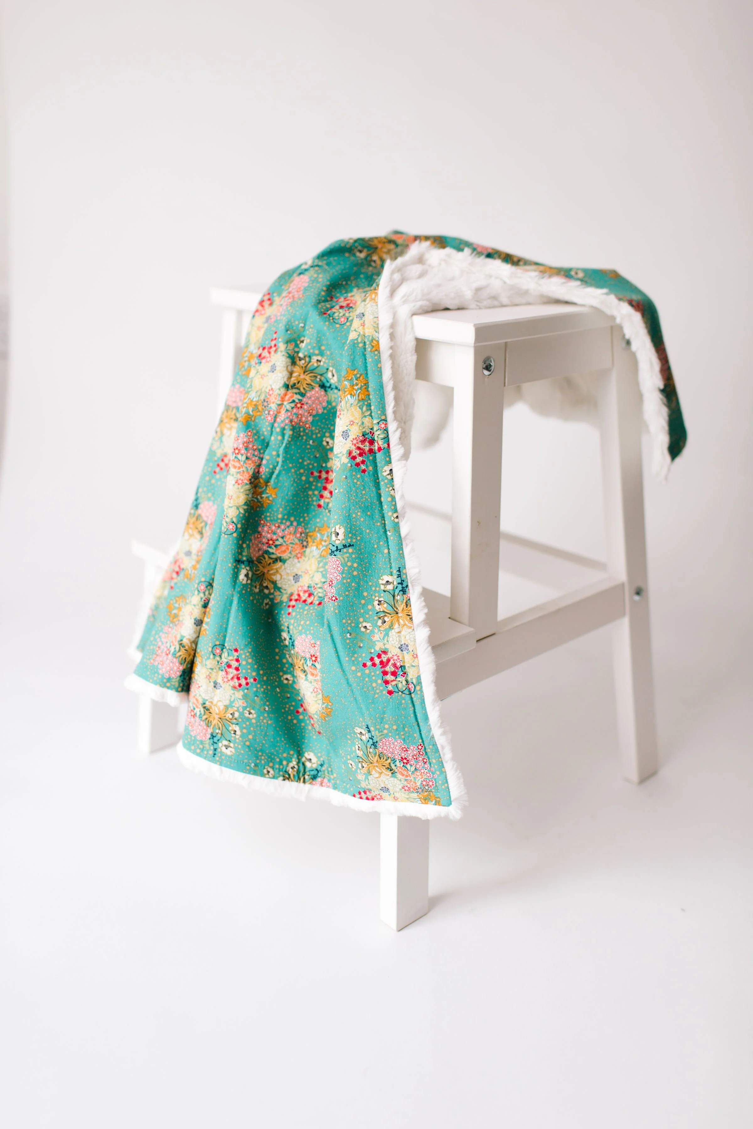 A white wooden step stool with a colorful, patterned fabric draped over it, featuring a floral design and a soft, white faux fur lining.