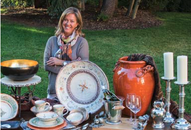 Smiling woman standing outdoors behind a table filled with various decorative and antique items like plates, bowls, a large orange vase, candles, and silverware.
