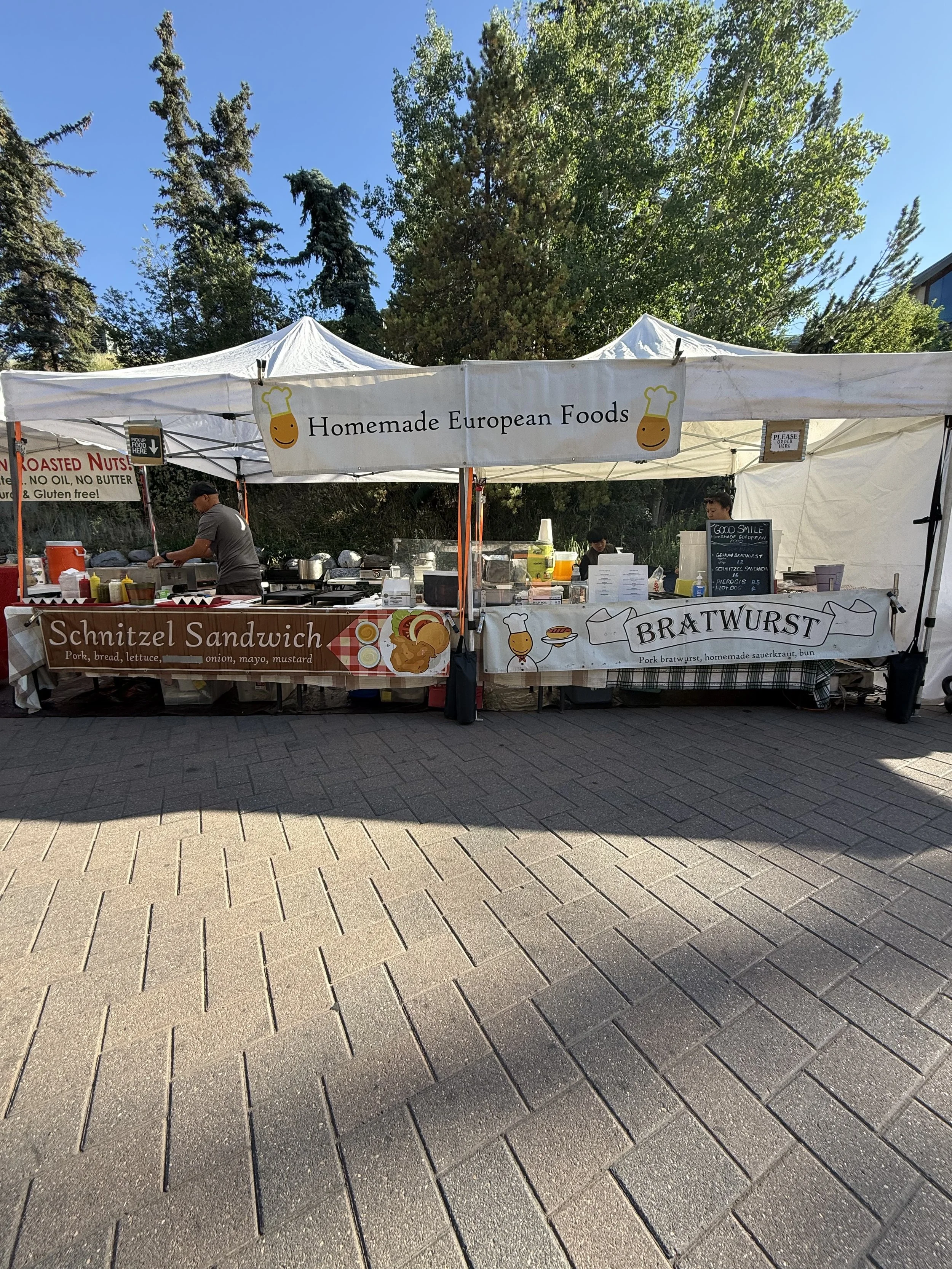 Outdoor food stand with signs for homemade European foods, schnitzel sandwiches, and bratwurst, with people preparing food under a white tent, trees in the background, and a paved surface in the foreground.