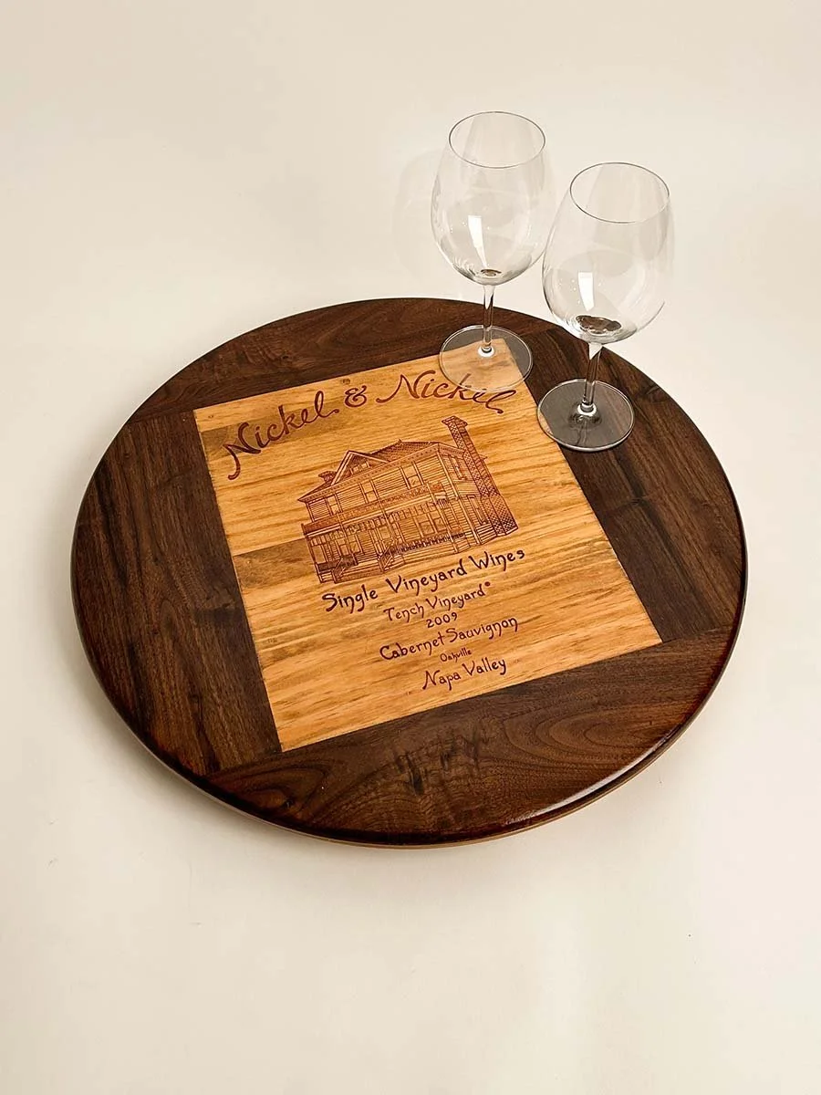 A round wooden tray with a square engraved wooden insert featuring a house illustration and text about Nickel & Nickle single vineyard wines from Napa Valley, with two empty wine glasses placed on top.