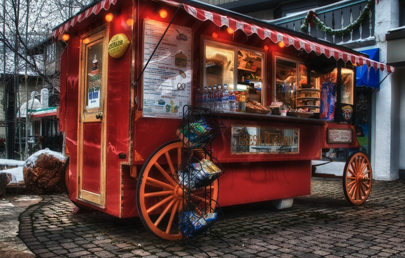 A red mobile food cart with orange wheels selling snacks like popcorn, with a menu and bottles of water displayed. The cart is decorated with string lights and is parked on cobblestone pavement outside, with snow and trees visible nearby.