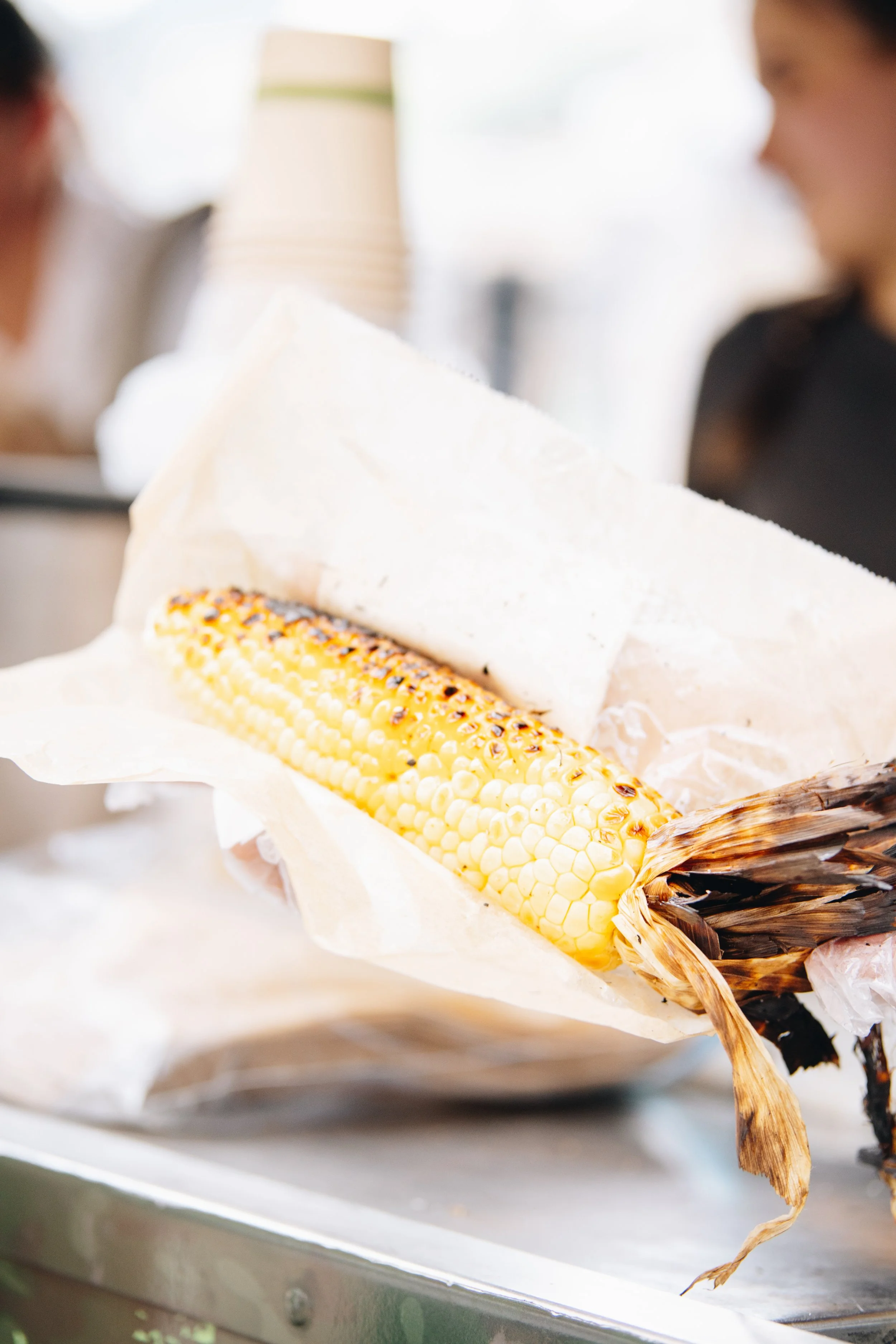 Elevated Elote A fresh ear of grilled corn on the cob with charred kernels, partially wrapped in white paper, held by a person with blurred background showing other people and a coffee cup.