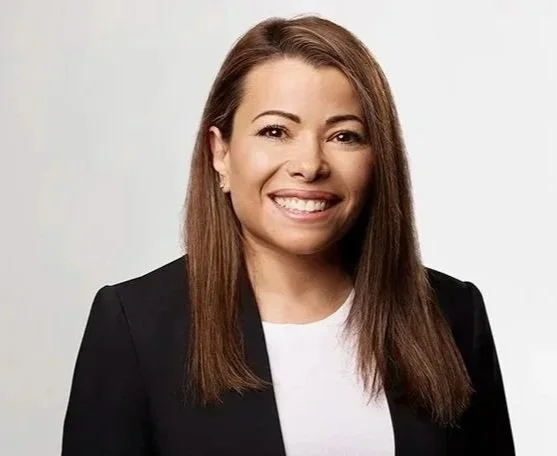 A woman with long brown hair smiling, wearing a black blazer over a white top, standing against a plain light gray background.