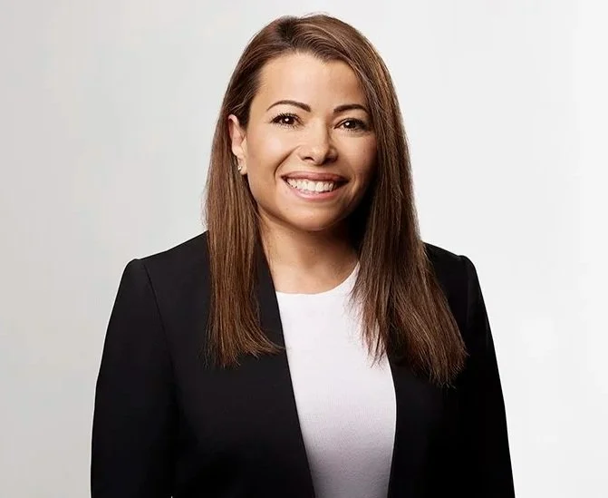 A woman with long brown hair smiling, wearing a black blazer over a white top, standing against a plain light gray background.