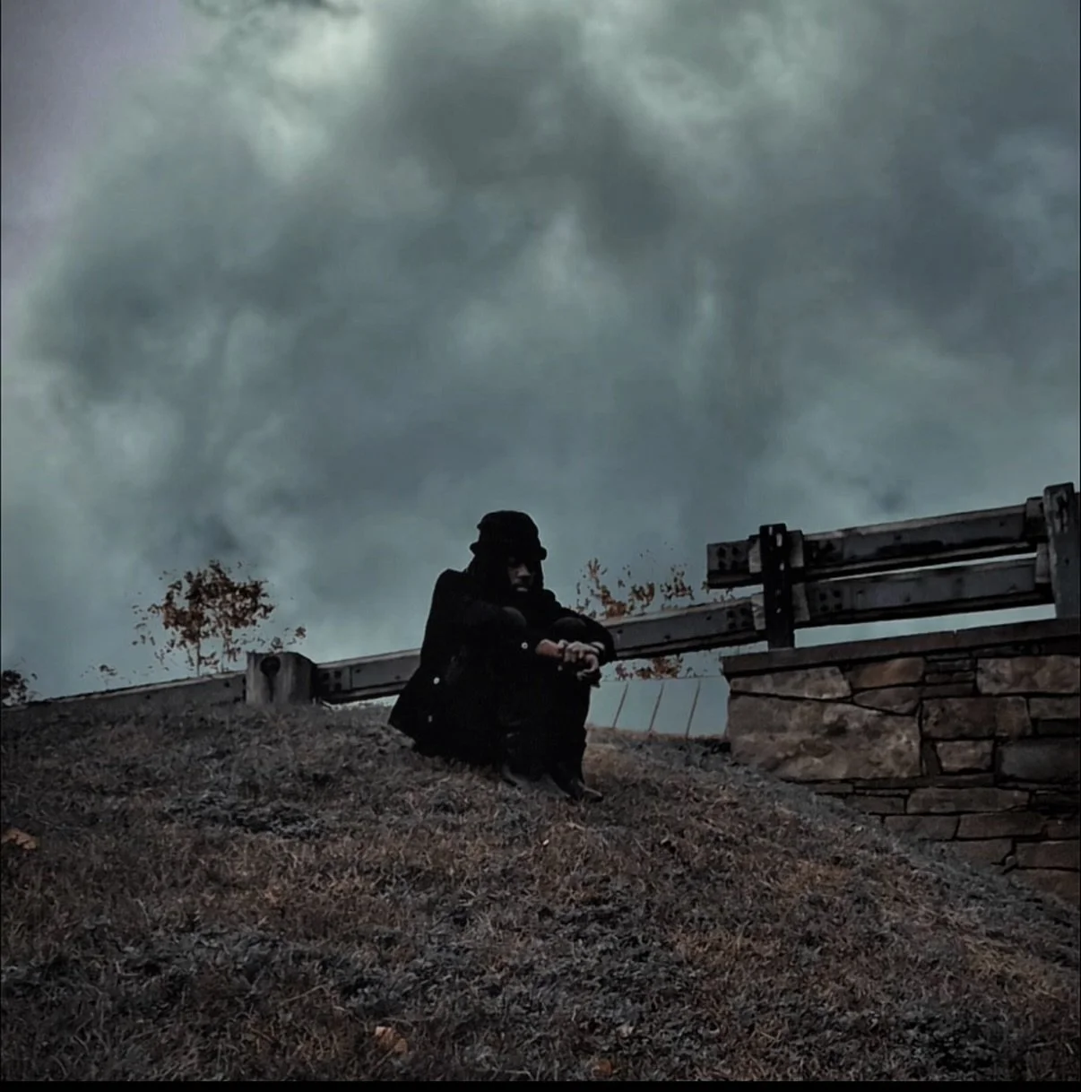 Person sitting on a grassy hillside near a stone wall and metal railing, with cloudy sky and a few trees in the background.