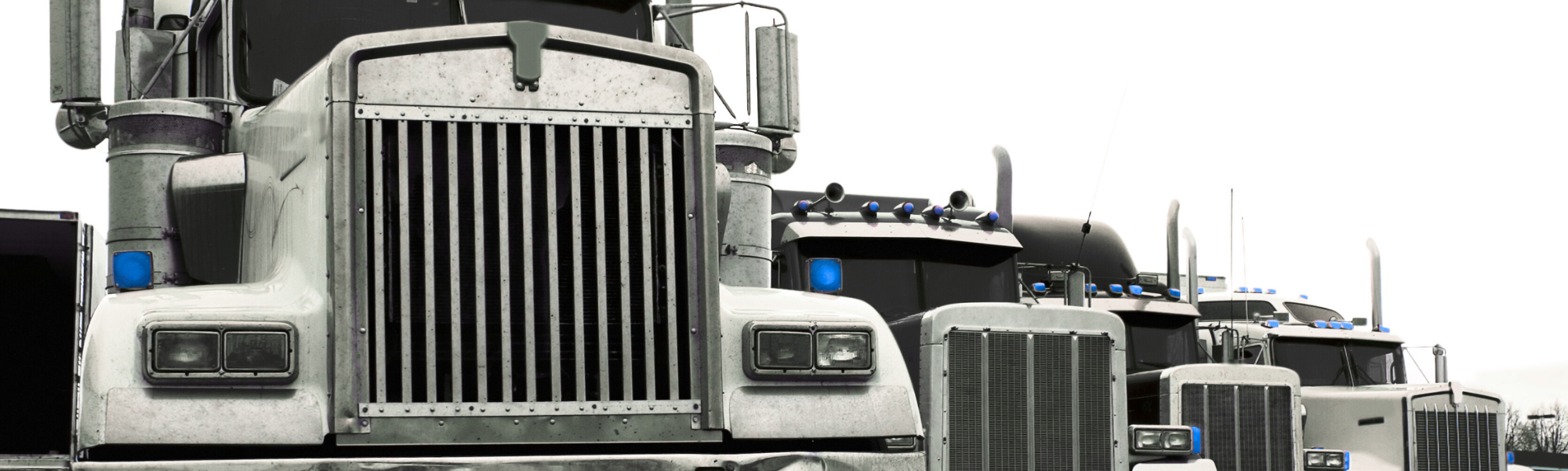 A lineup of large semi-trucks parked side by side outdoors, showing front views with prominent grills, headlights, and blue lights on top of the cabins.