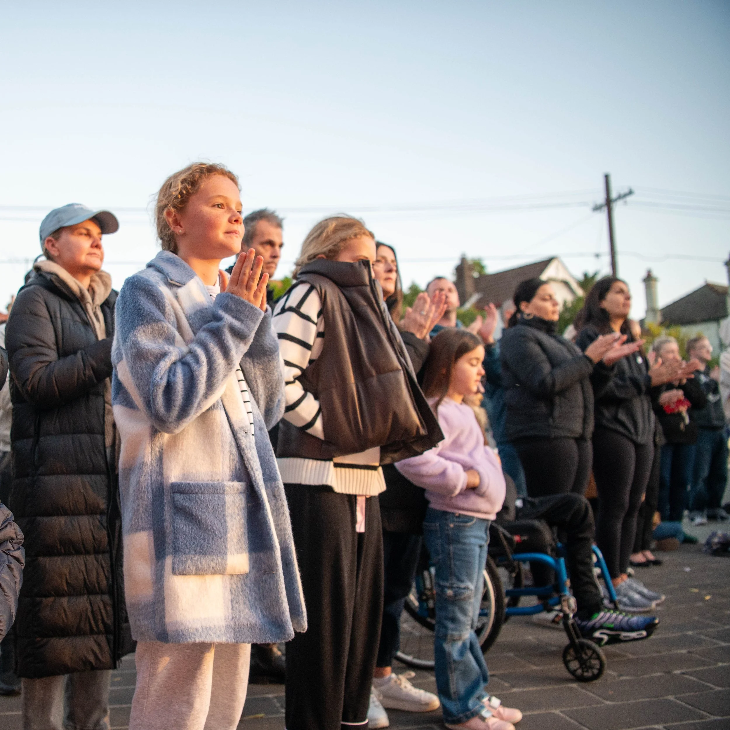 A group of people, including children and adults, standing outdoors with some holding their hands together in prayer or reflection during what appears to be a gathering or vigil.