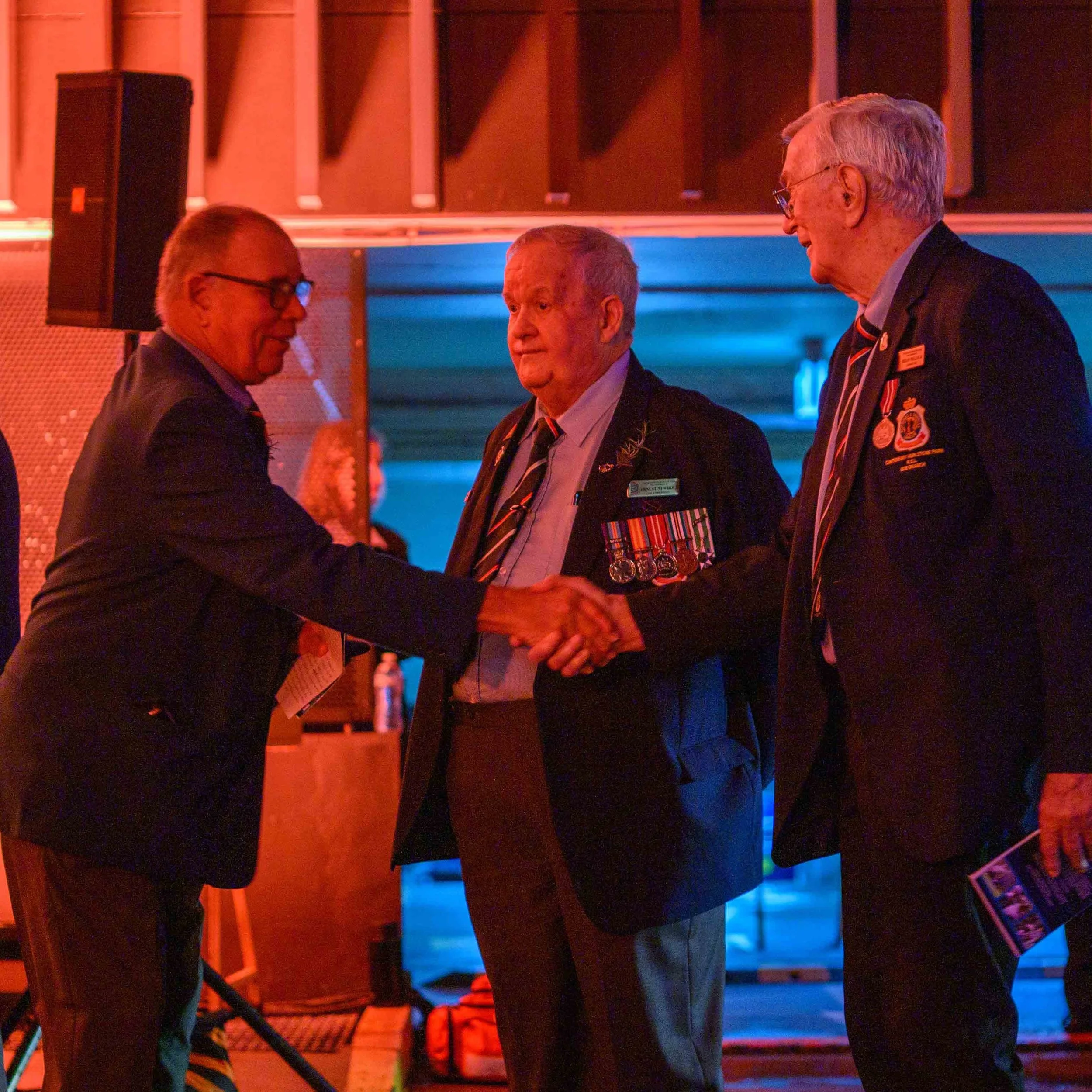Three men, one in military medals, shake hands at an indoor event with colorful lighting.