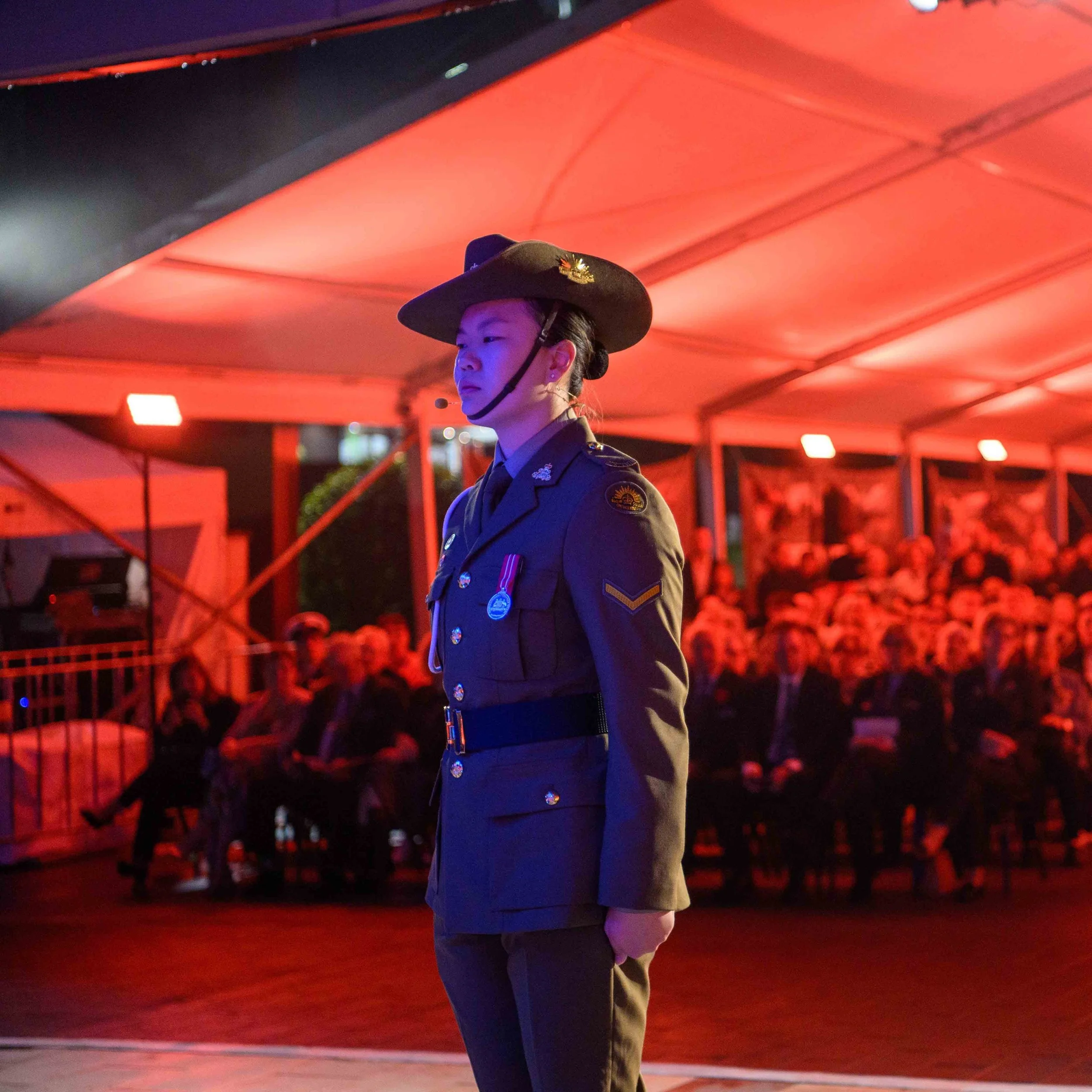 A female soldier in uniform standing at attention during a formal event under red lighting, with a seated audience in the background.