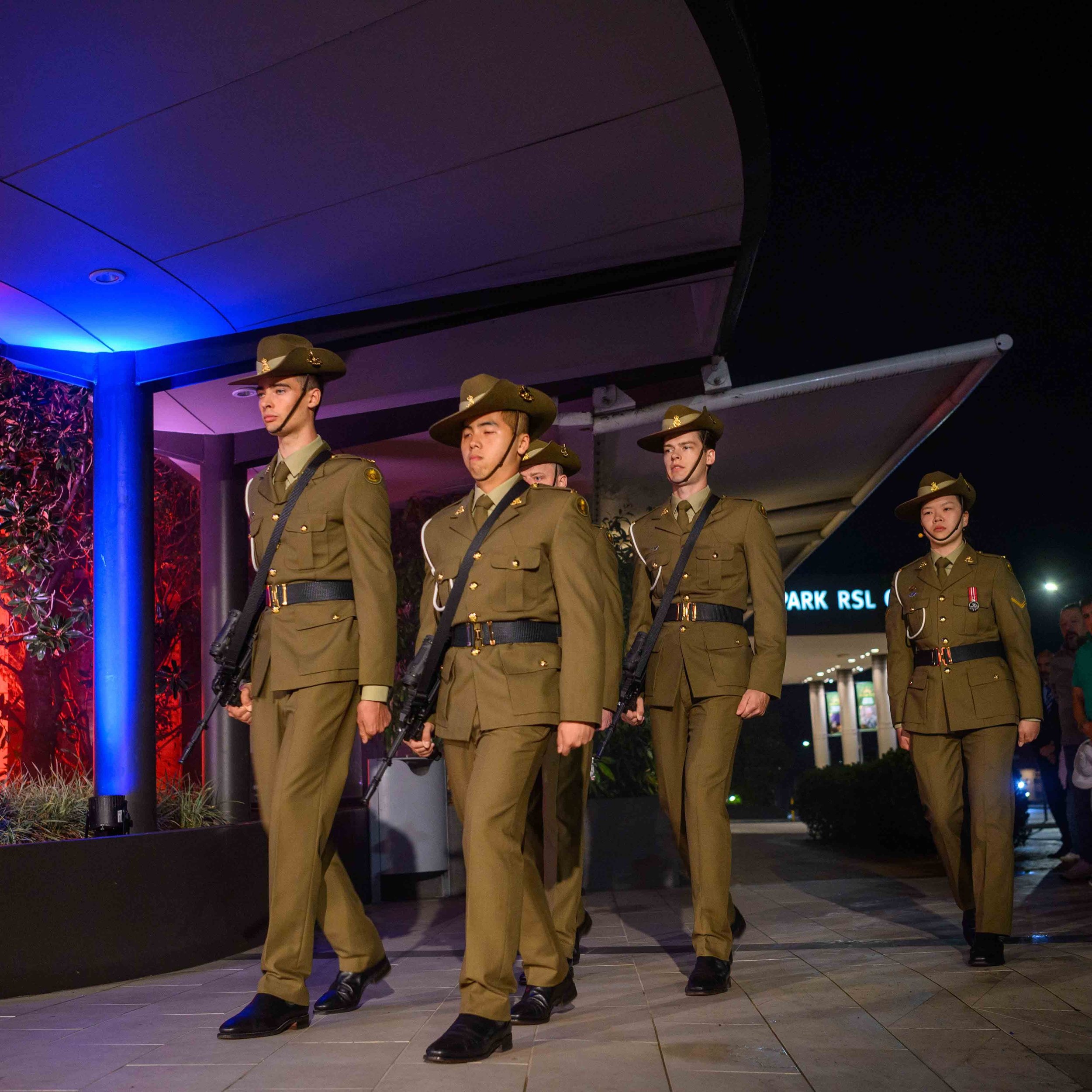 Group of five soldiers in ACU dress uniforms and campaign hats, marching in formation outside a building at night.