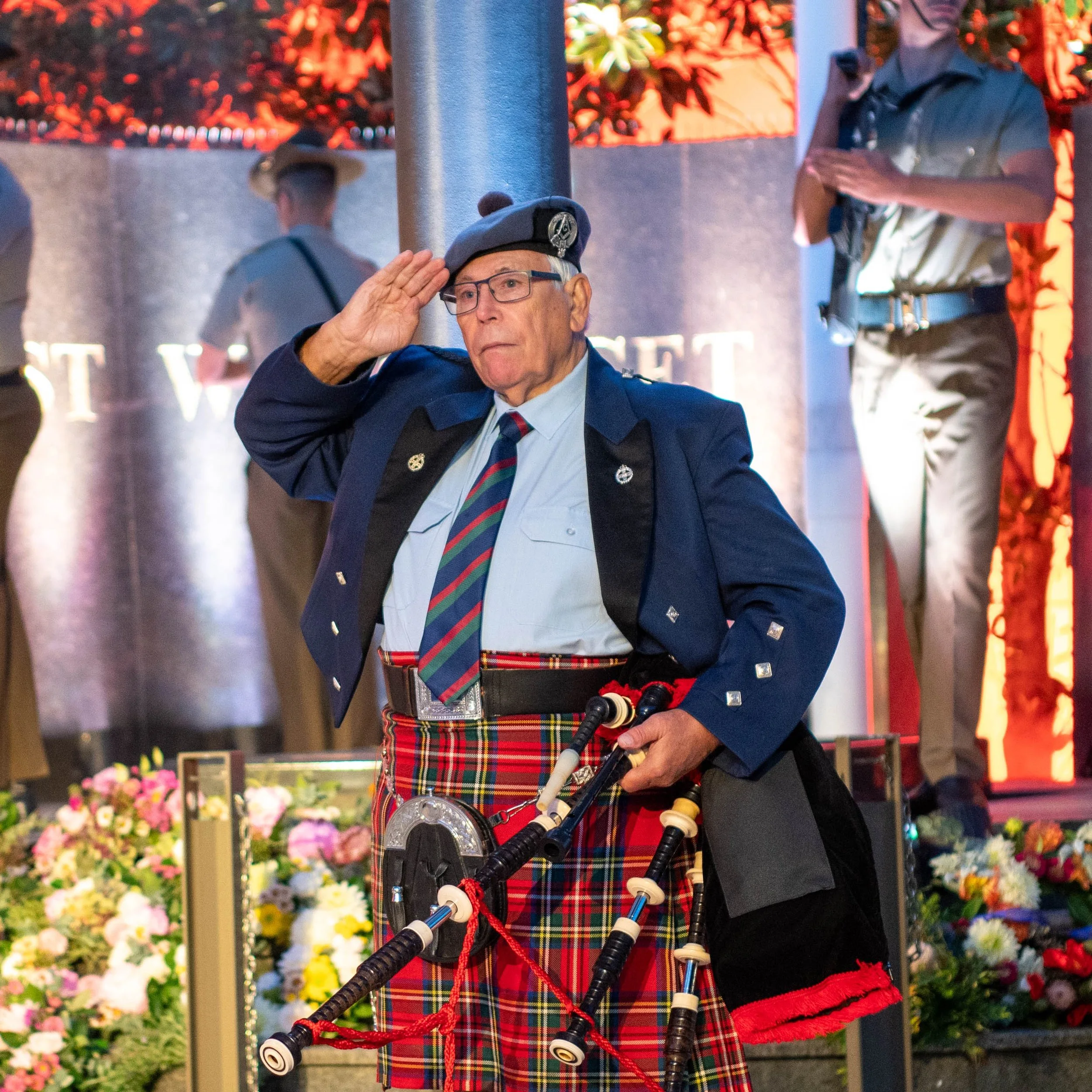 An elderly man in a Scottish-style kilt and uniform salutes during a formal event. He is holding a set of bagpipes, standing in front of a floral arrangement with people in uniforms and a stage backdrop.