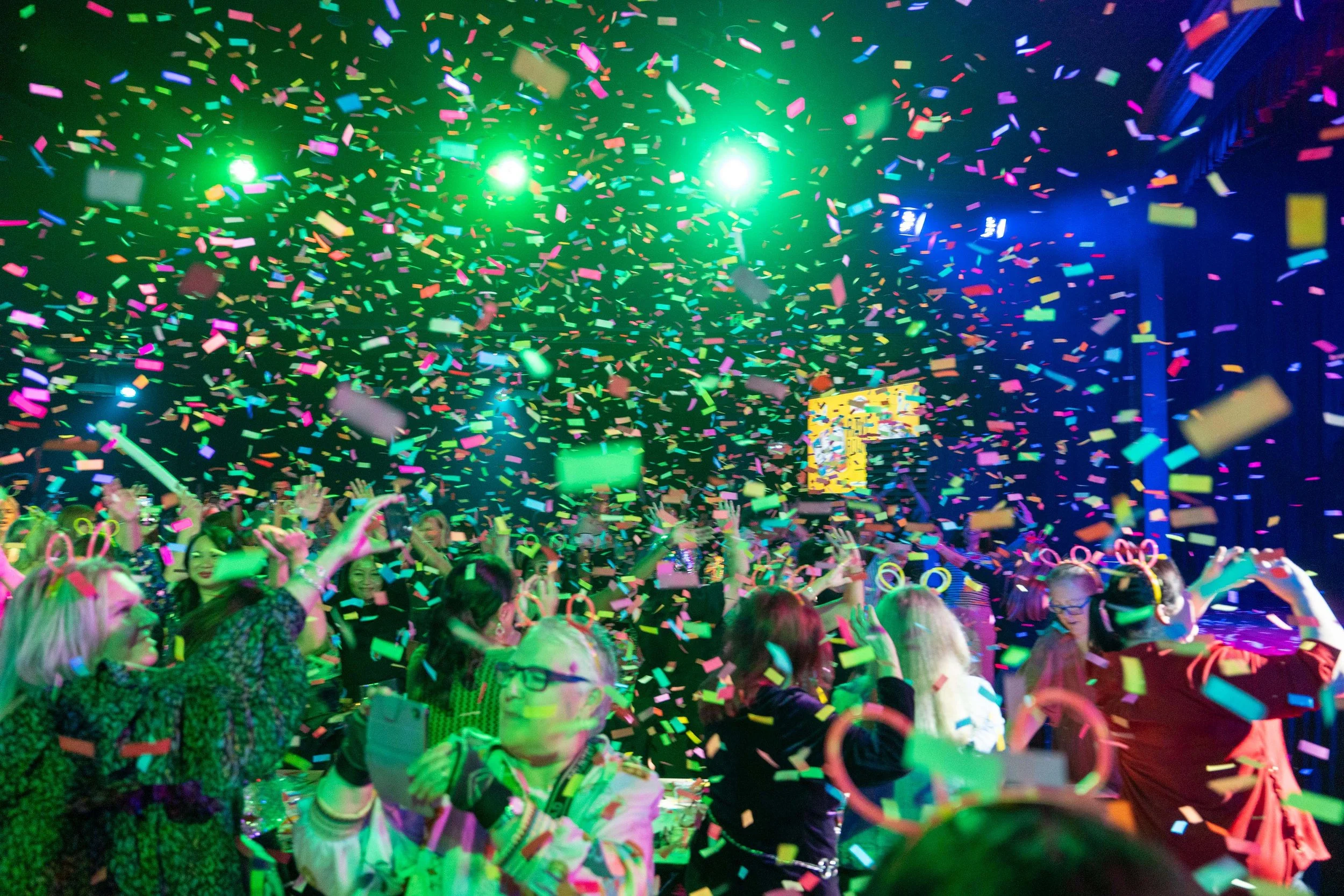 Crowd celebrating with colorful confetti and bright green lights at an indoor event or party.