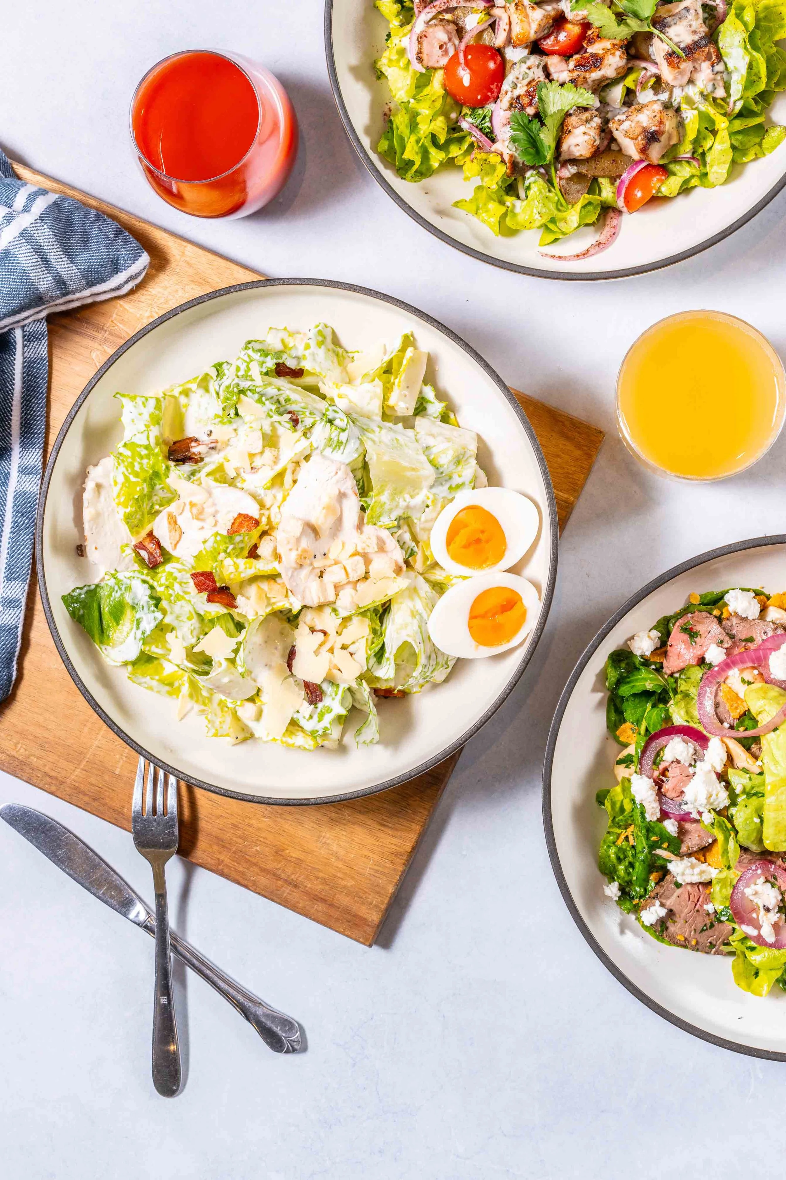 Salad plates with lettuce, cherry tomatoes, eggs, cheese, and grilled chicken; glasses of red and orange beverages; a blue and white napkin; a fork and knife; and a wooden cutting board.