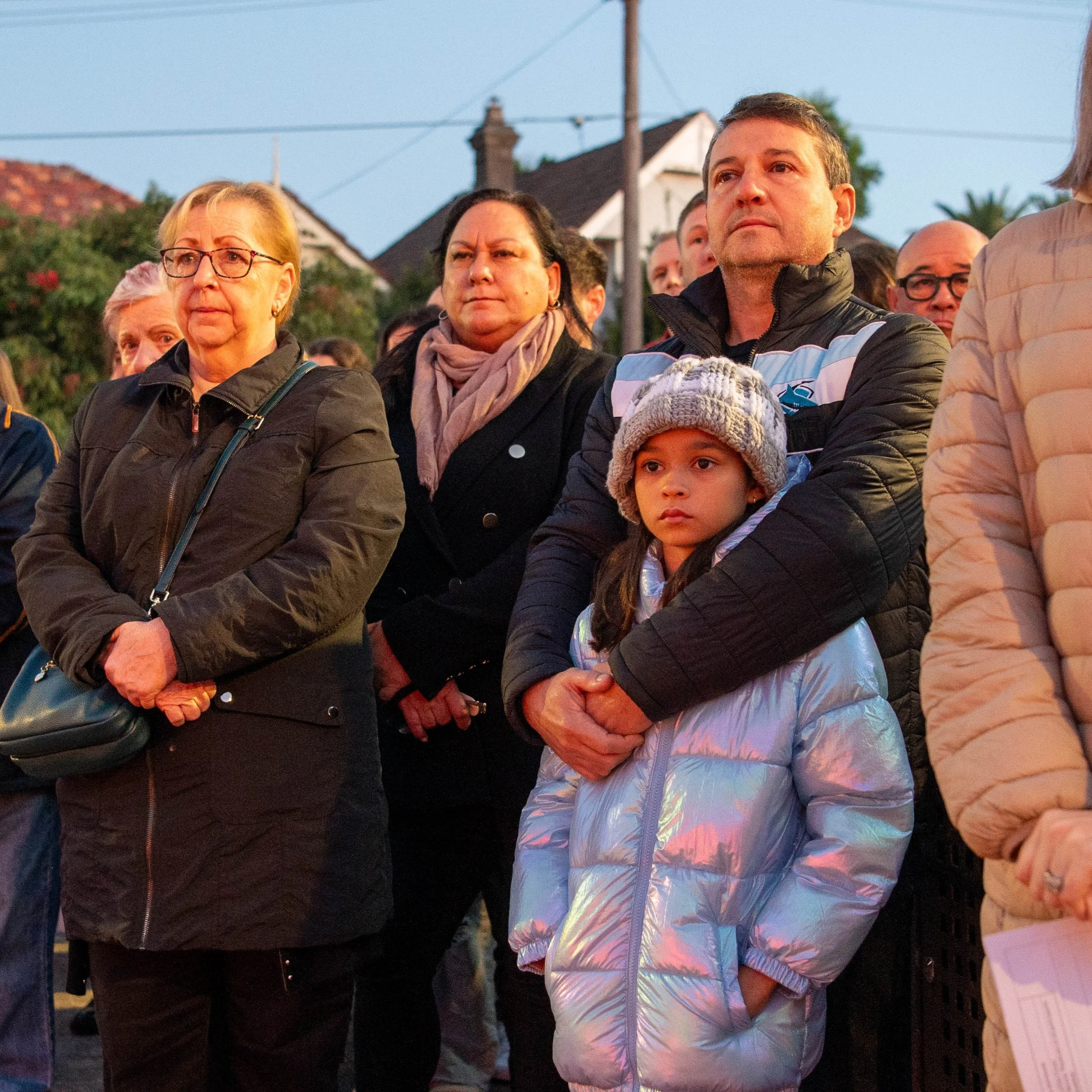 A group of people standing outdoors during the evening, watching solemnly. The scene includes a woman with glasses, a woman with a scarf, a man holding a young girl wearing a gray knit hat and a shiny jacket, surrounded by others in jackets and coats.