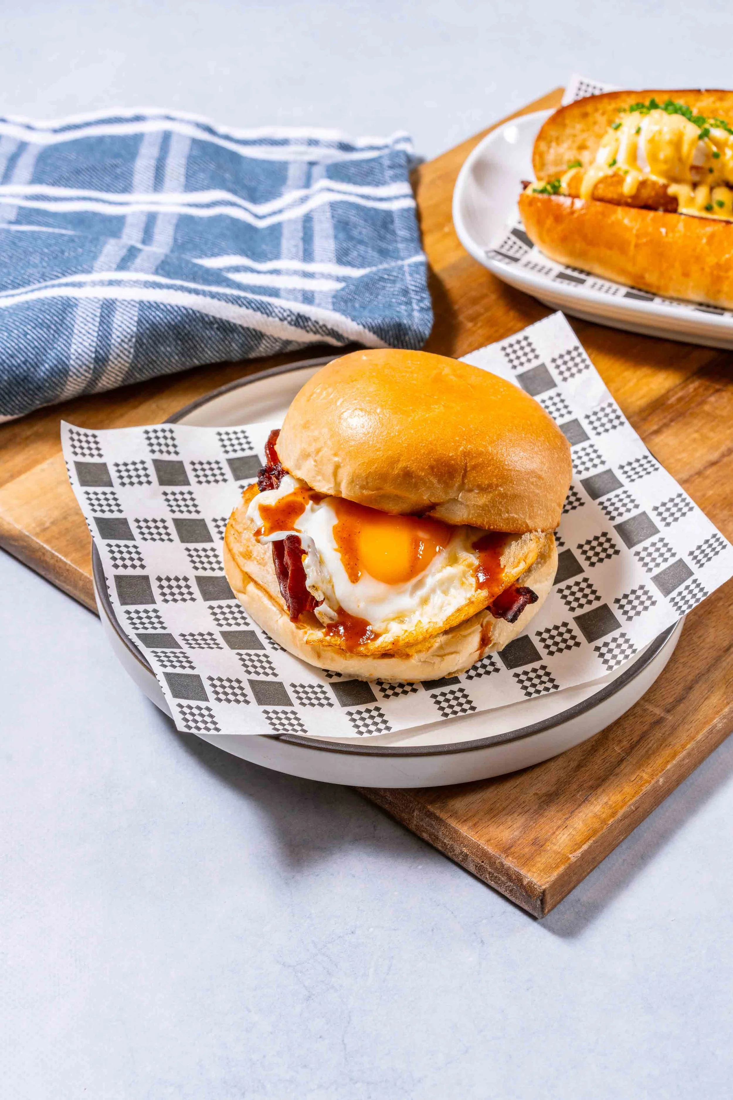 Close-up of a breakfast sandwich with a fried egg, bacon, on a bun, served on a plate with checkered paper, with an egg salad sandwich in the background on a plate.