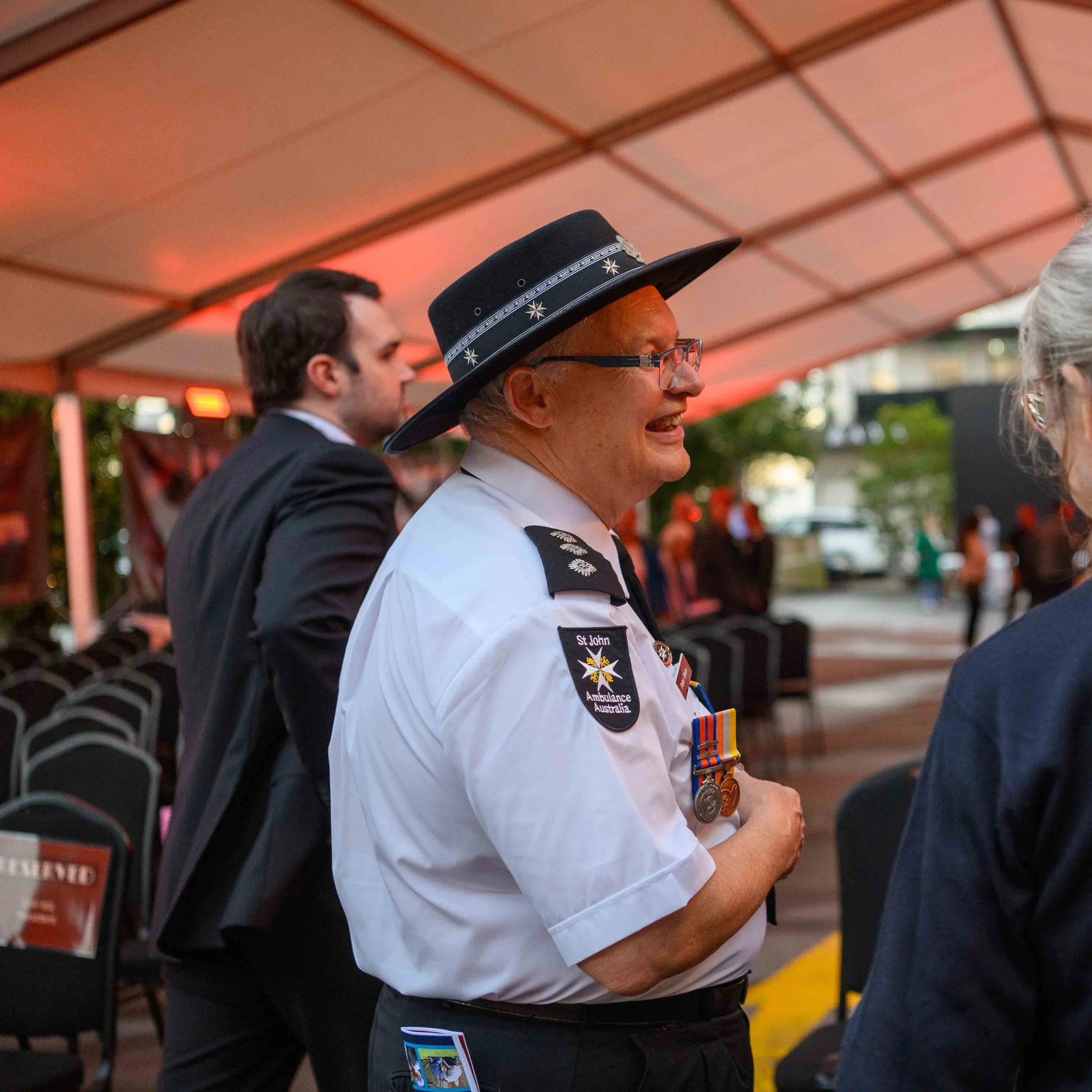 An elderly man in a white uniform with medals and shoulder insignia, wearing a black hat with a decorative band, is smiling and conversing with a woman with gray hair. A younger man in a black suit stands behind him. The background shows chairs and an outdoor event setting with a tent canopy and other people.