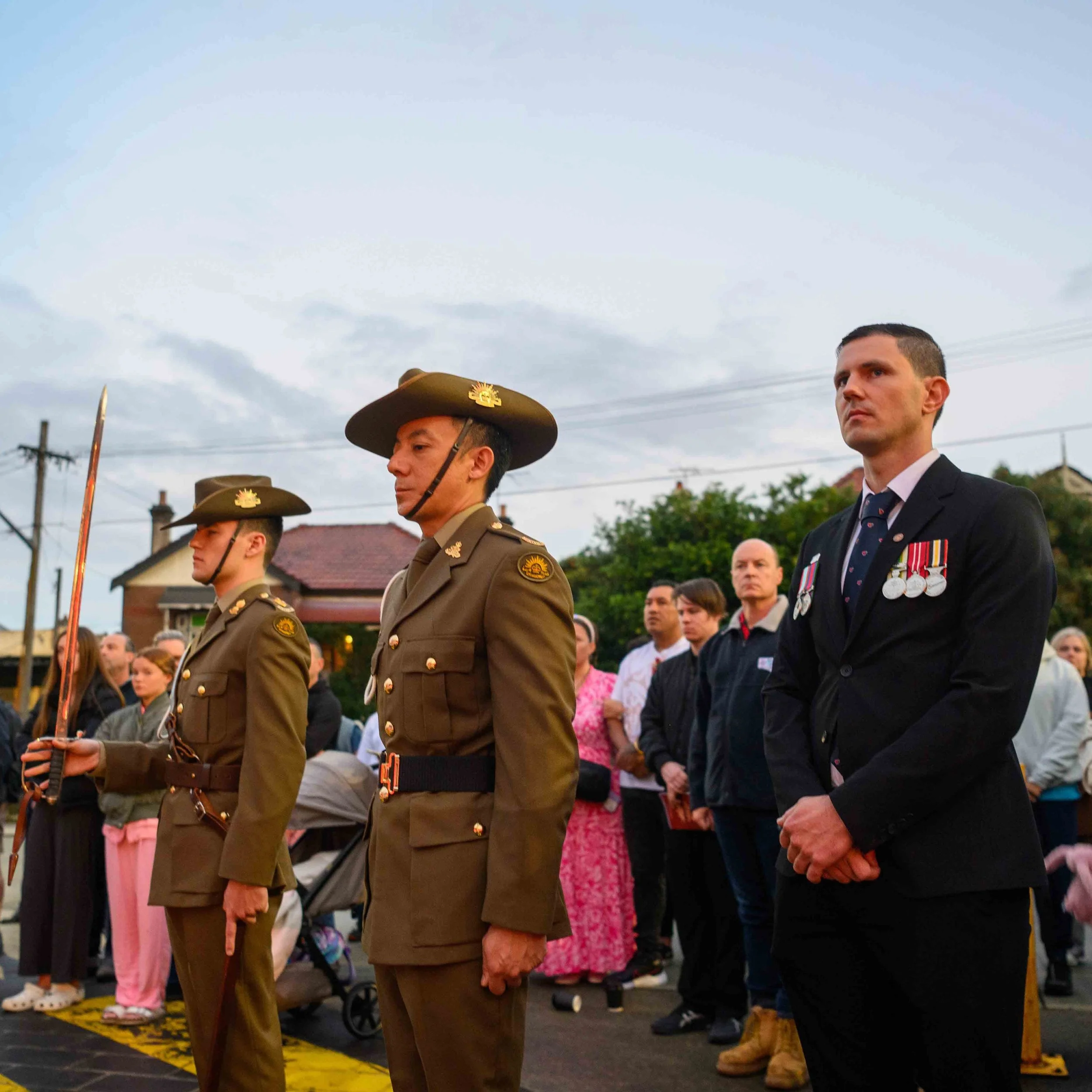 Military personnel and civilians attending a memorial or ceremony, standing solemnly outdoors, with houses and trees in the background.