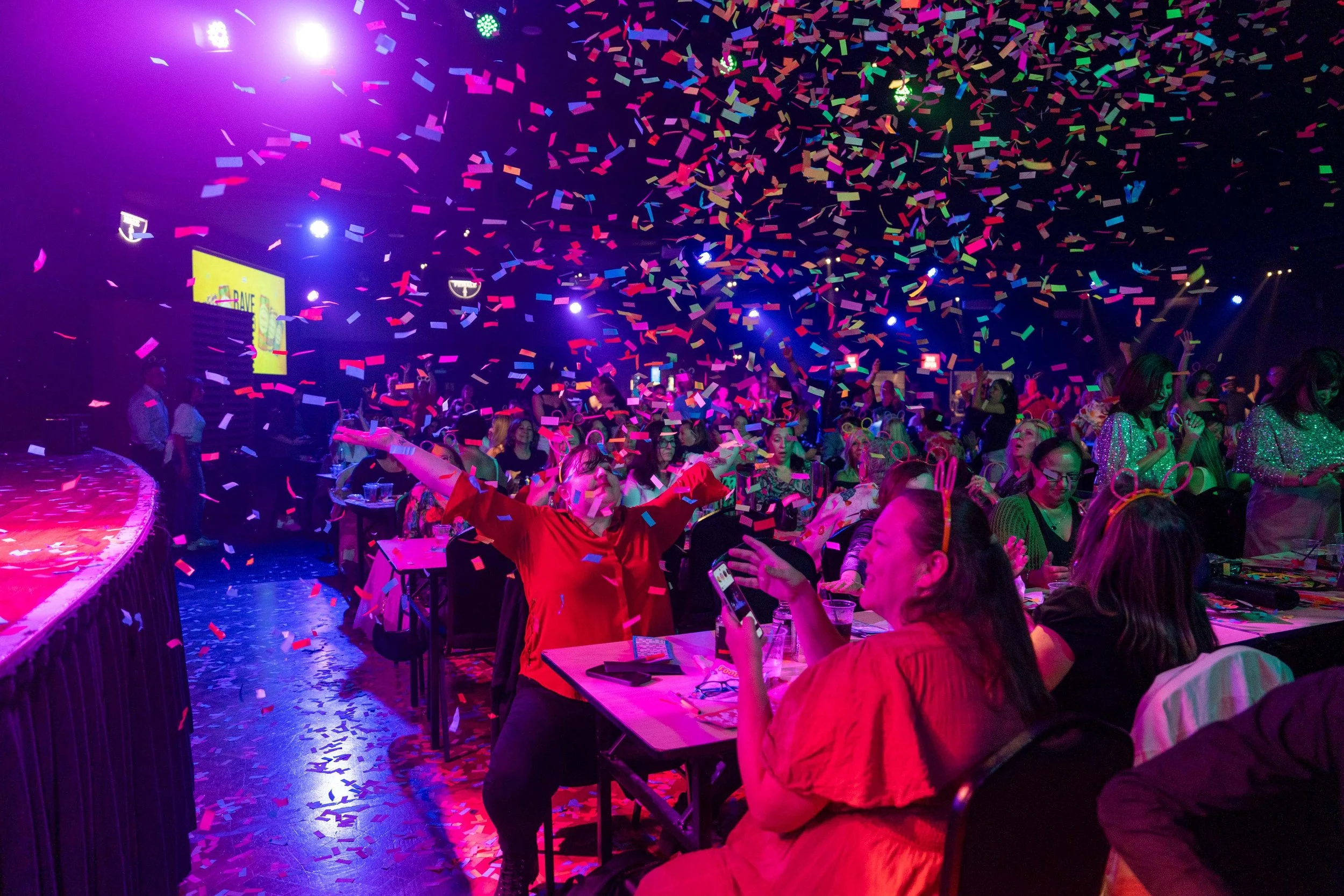 Crowd of people celebrating at an event with colorful confetti and stage lights, some taking photos, in a festive indoor setting.