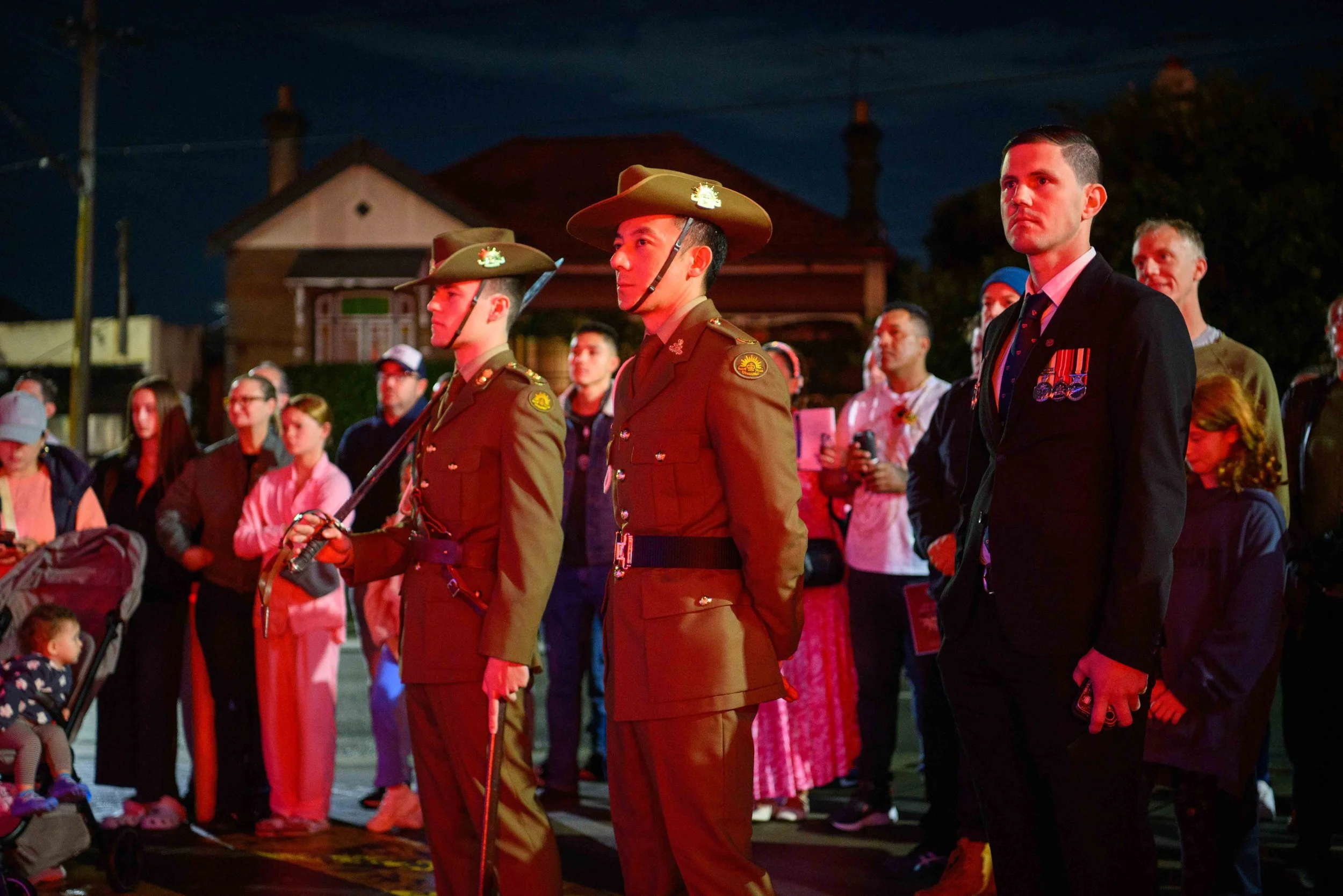 Nighttime gathering of military personnel and civilians, some in formal attire with medals, and others in casual clothing, standing solemnly outside a residential area.