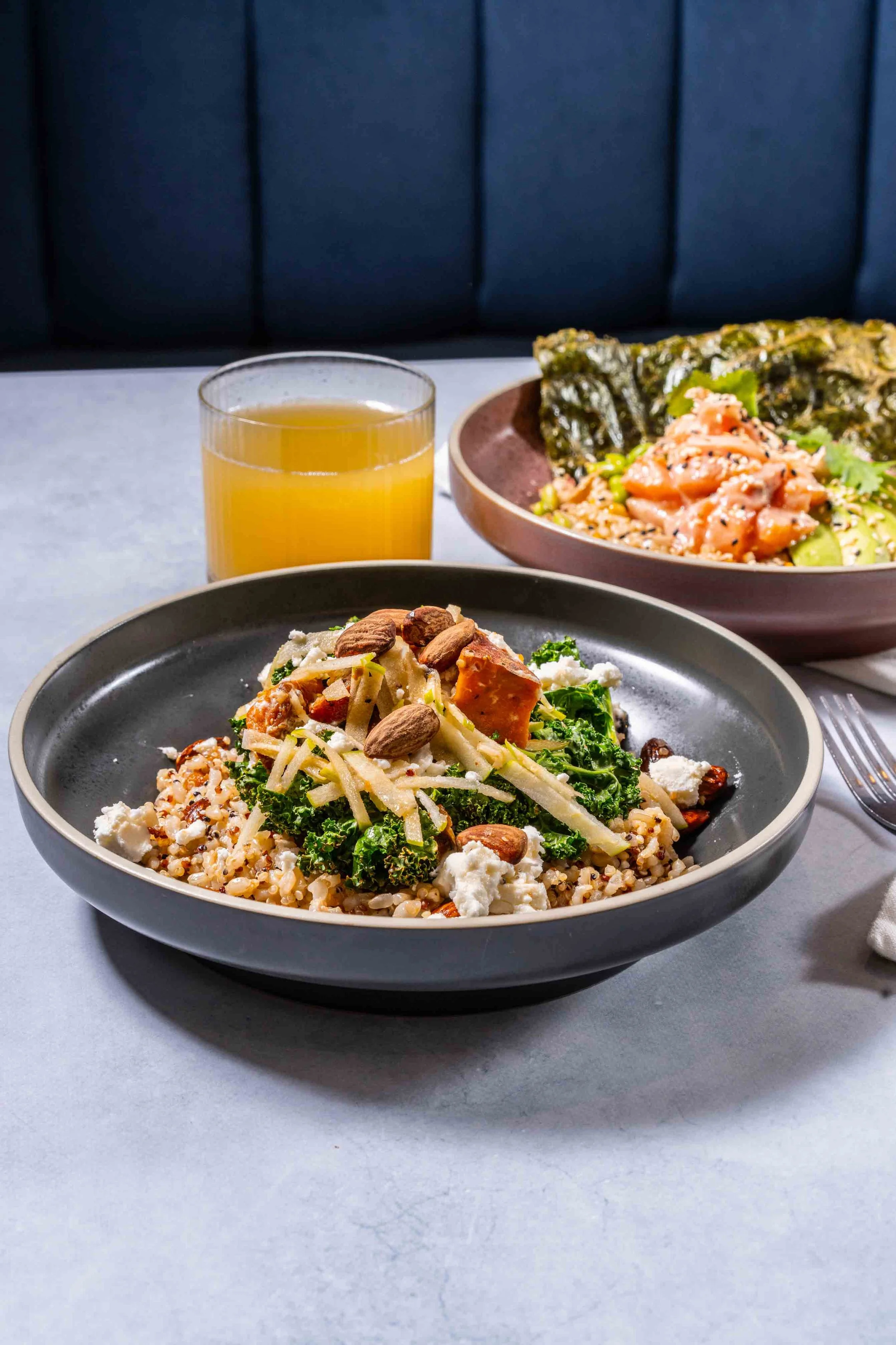 A bowl of grain salad with kale, cheese, almonds, and other vegetables, served with a glass of orange juice and another bowl of rice and seafood in the background.