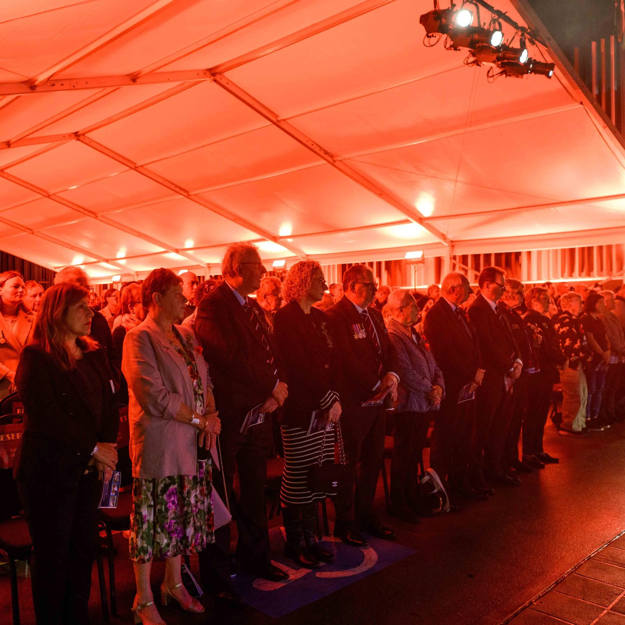 A group of people standing in a line under a large, red-lit tent at an event, some dressed in formal attire.