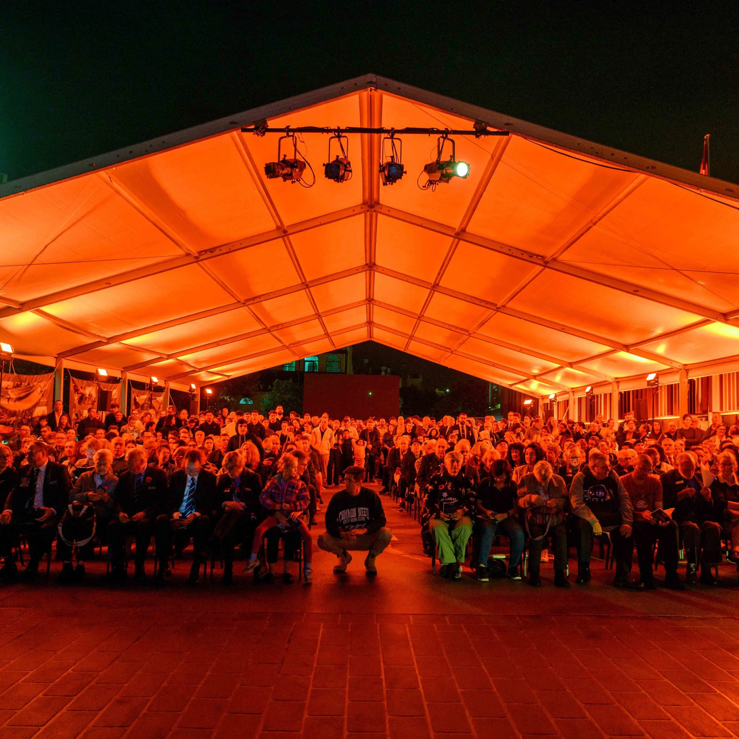 A large outdoor event taking place under a big orange tent at night with many seated and standing people, and stage lighting fixtures overhead.