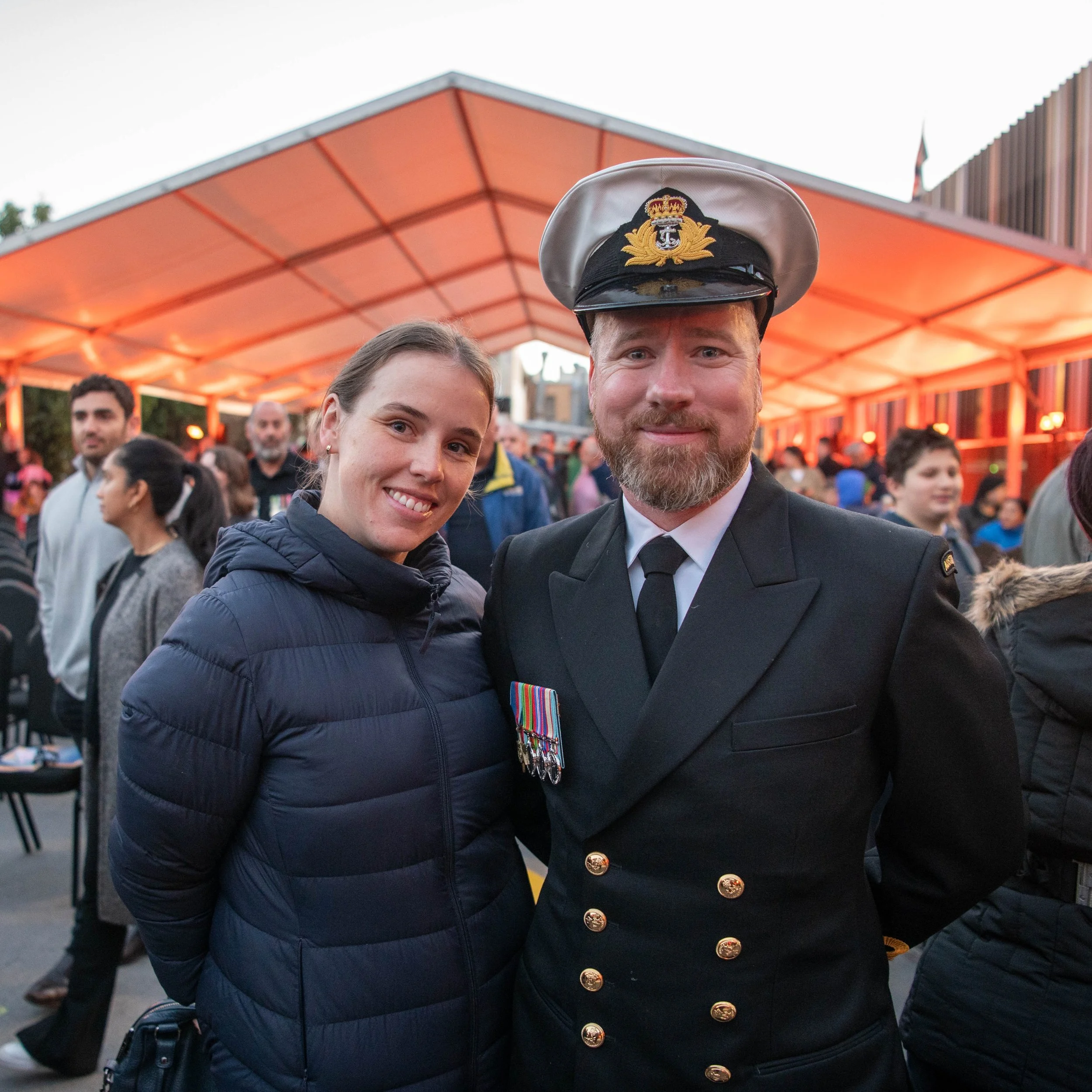 A woman and a man in a United States Navy officer uniform with medals, standing close together and smiling at the camera at an outdoor event with a crowd and orange tent in background.
