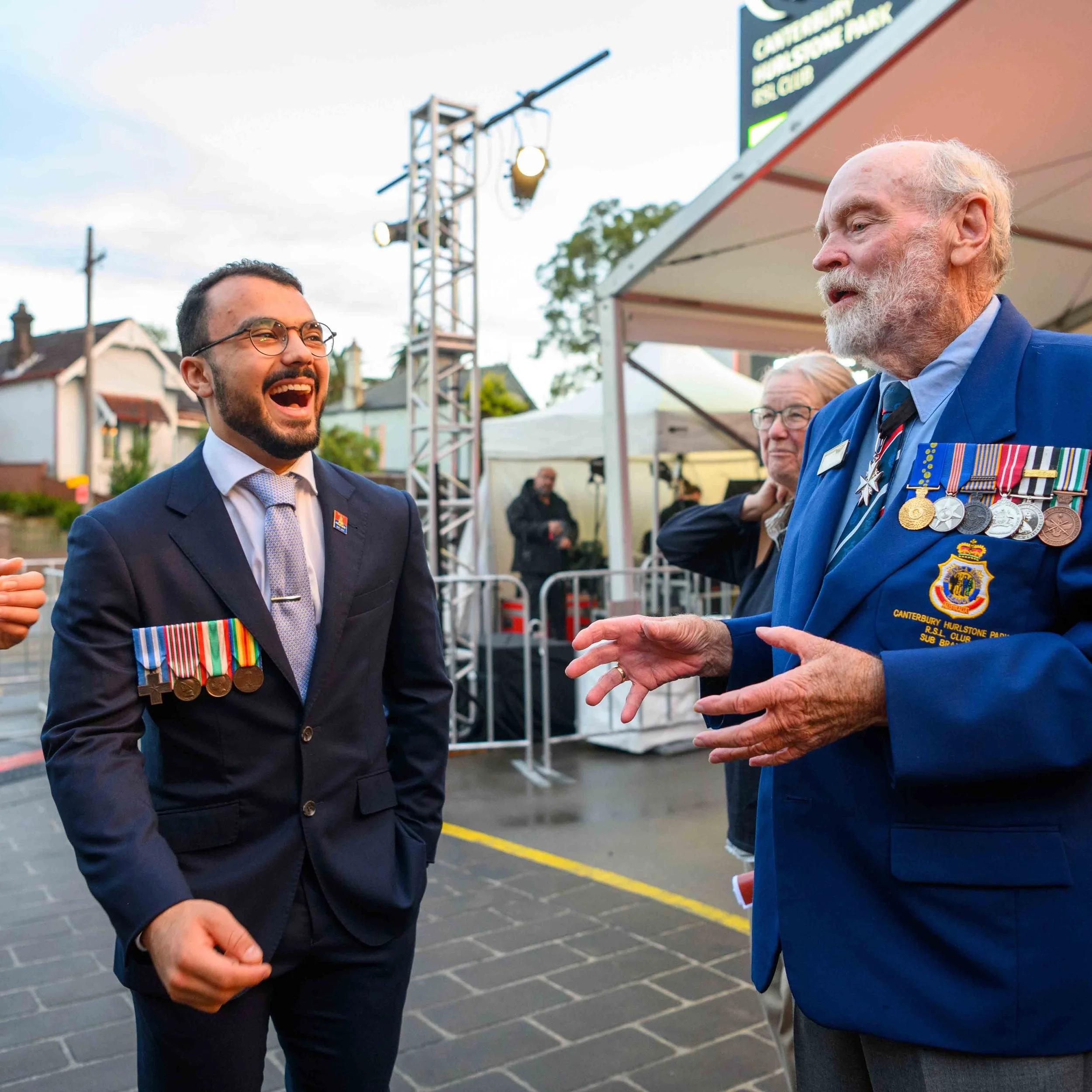 Two men in military medals and formal attire engaged in conversation at an outdoor event, with a woman and a crowd in the background.