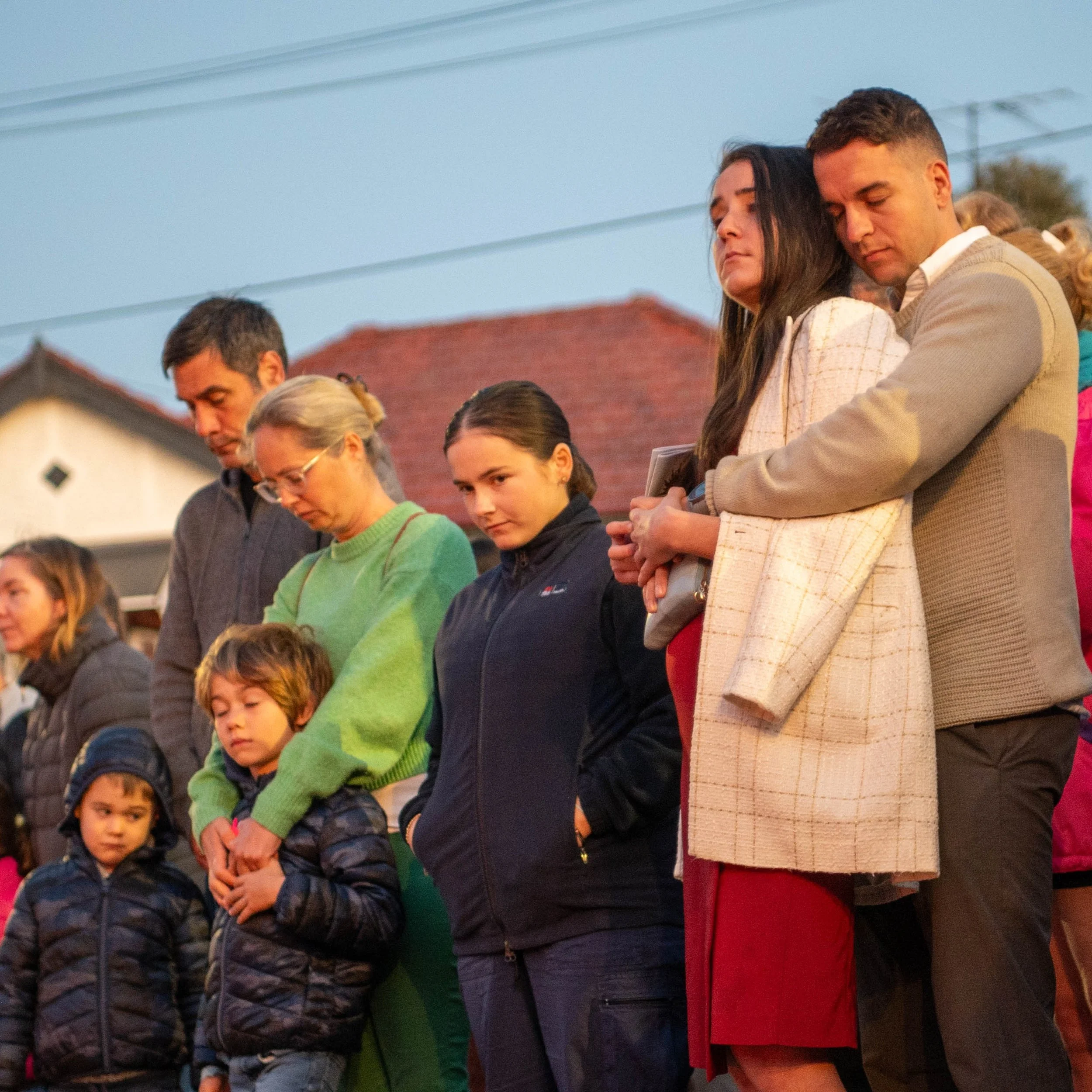 A group of people standing outdoors during the evening, with some people with eyes closed, seemingly in prayer or reflection.