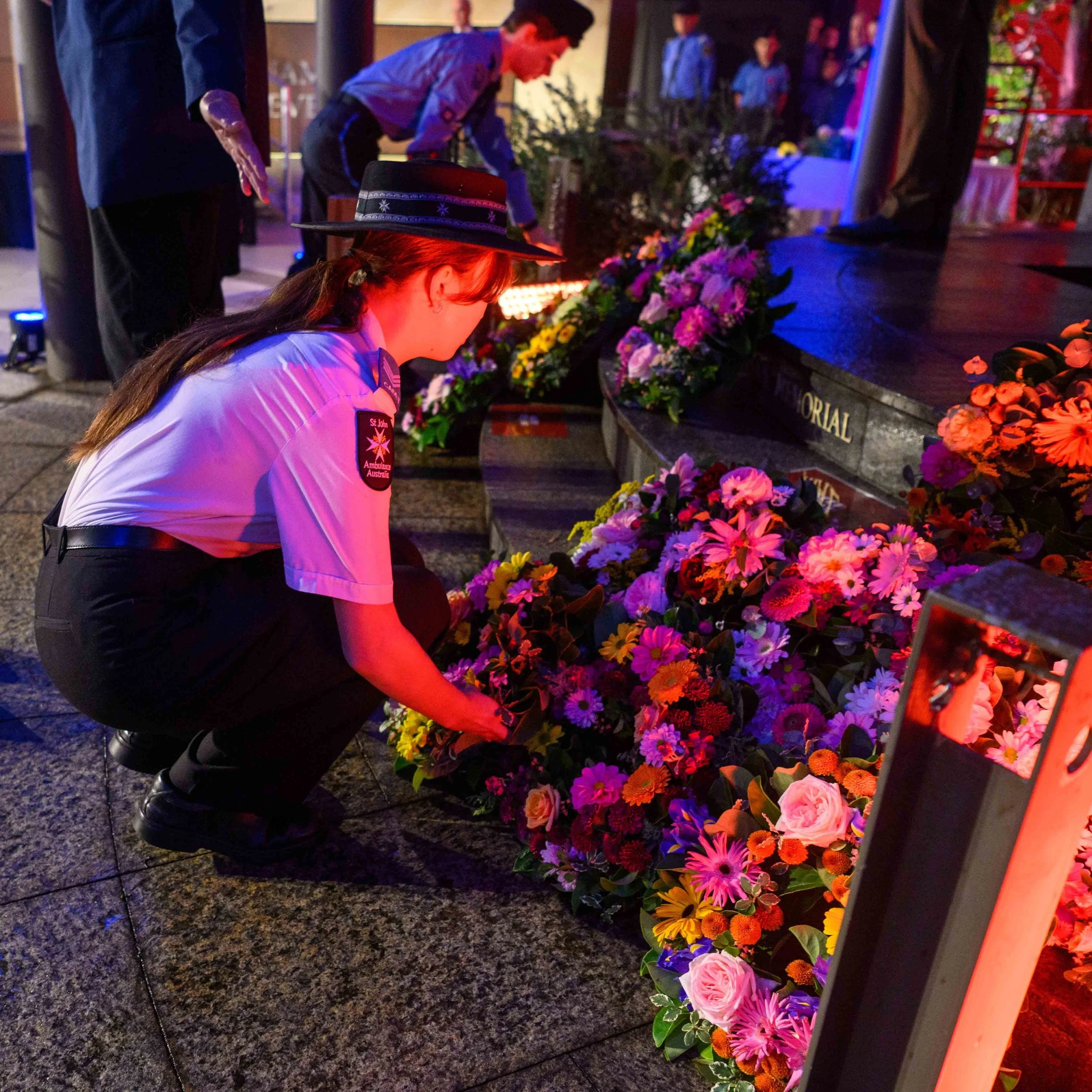 A uniformed woman placing flowers at a memorial, with other people also participating in the memorial event, illuminated by colorful lights.