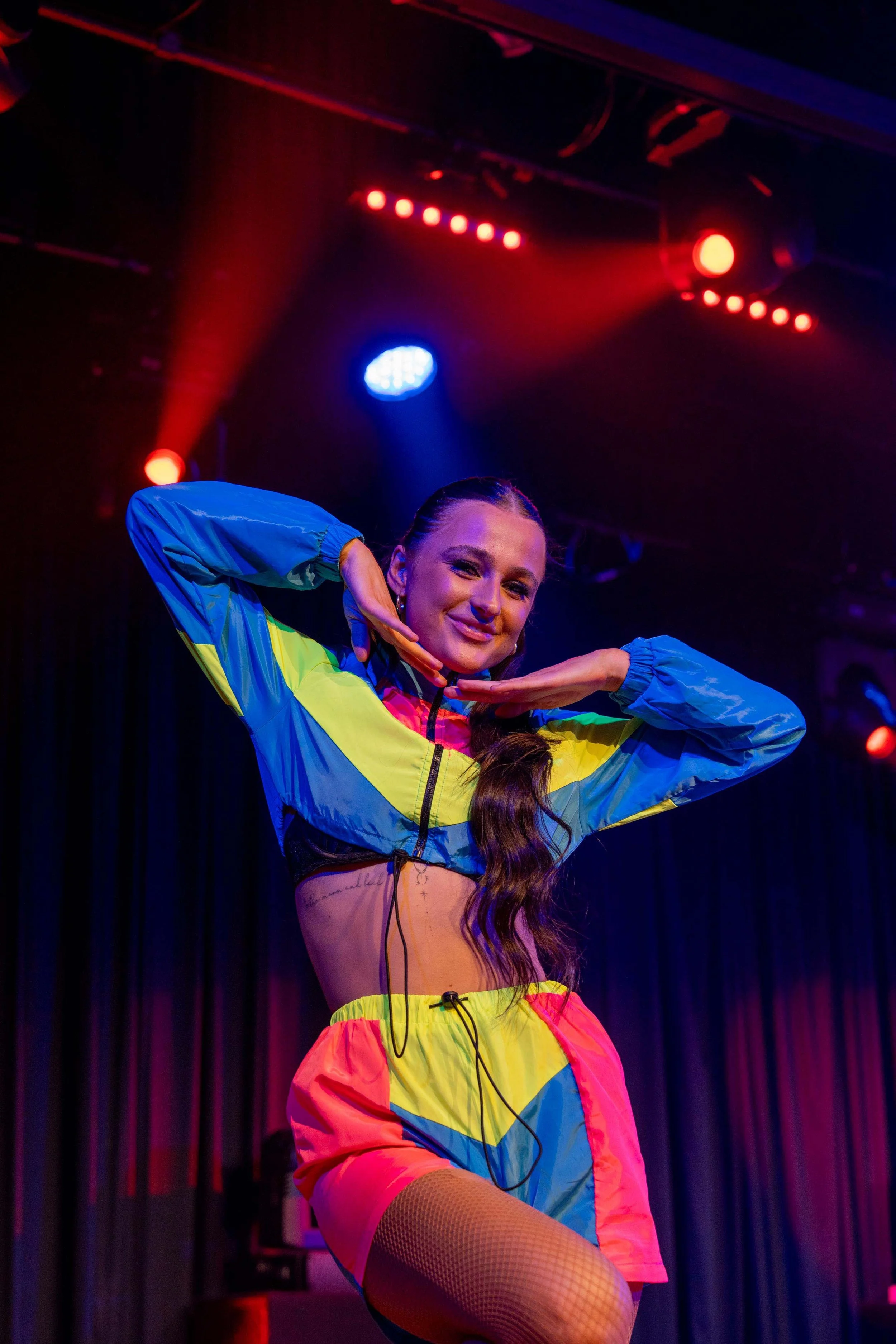 A woman dancing on stage under colorful red, blue, and purple stage lights, wearing a vibrant color-blocked athletic outfit including a cropped jacket and shorts.