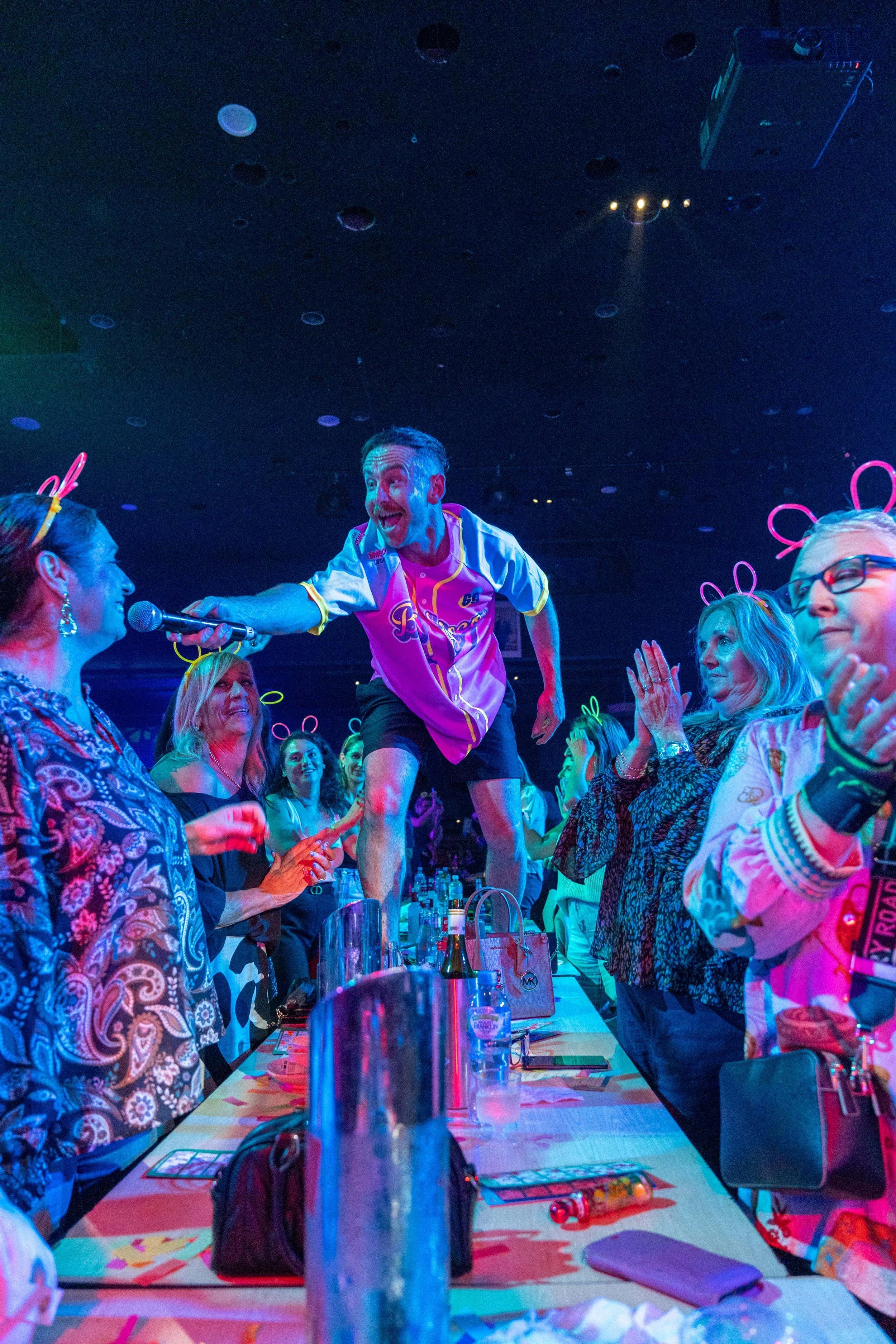 A lively event with a man in a pink sports jersey standing on a table, handing a microphone to a woman, surrounded by people clapping and smiling, with neon-colored lights and a dark ceiling.