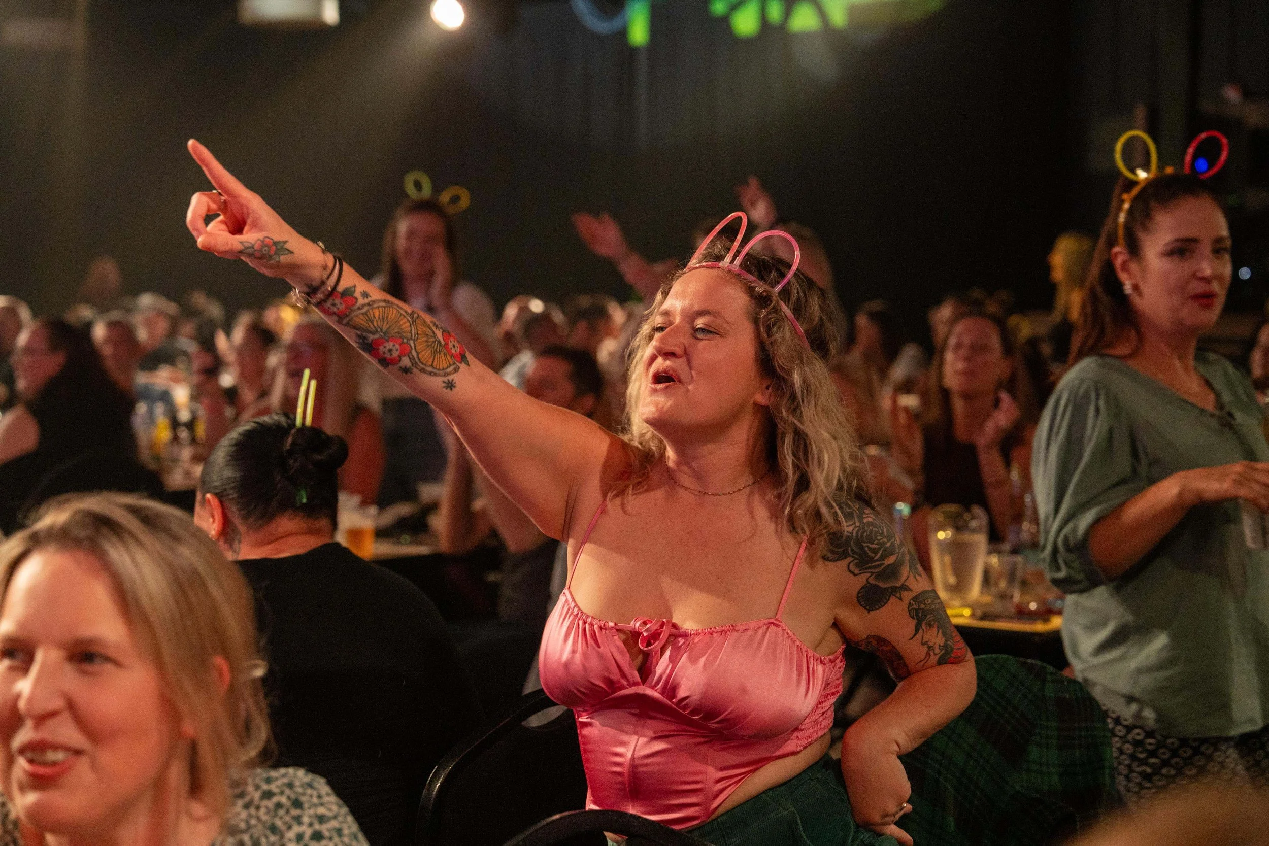 A woman in a pink satin top with tattoos on her arms and a pink headband in her hair is pointing and singing at a lively party or concert, with many other people in the background wearing glow headbands and enjoying the event.