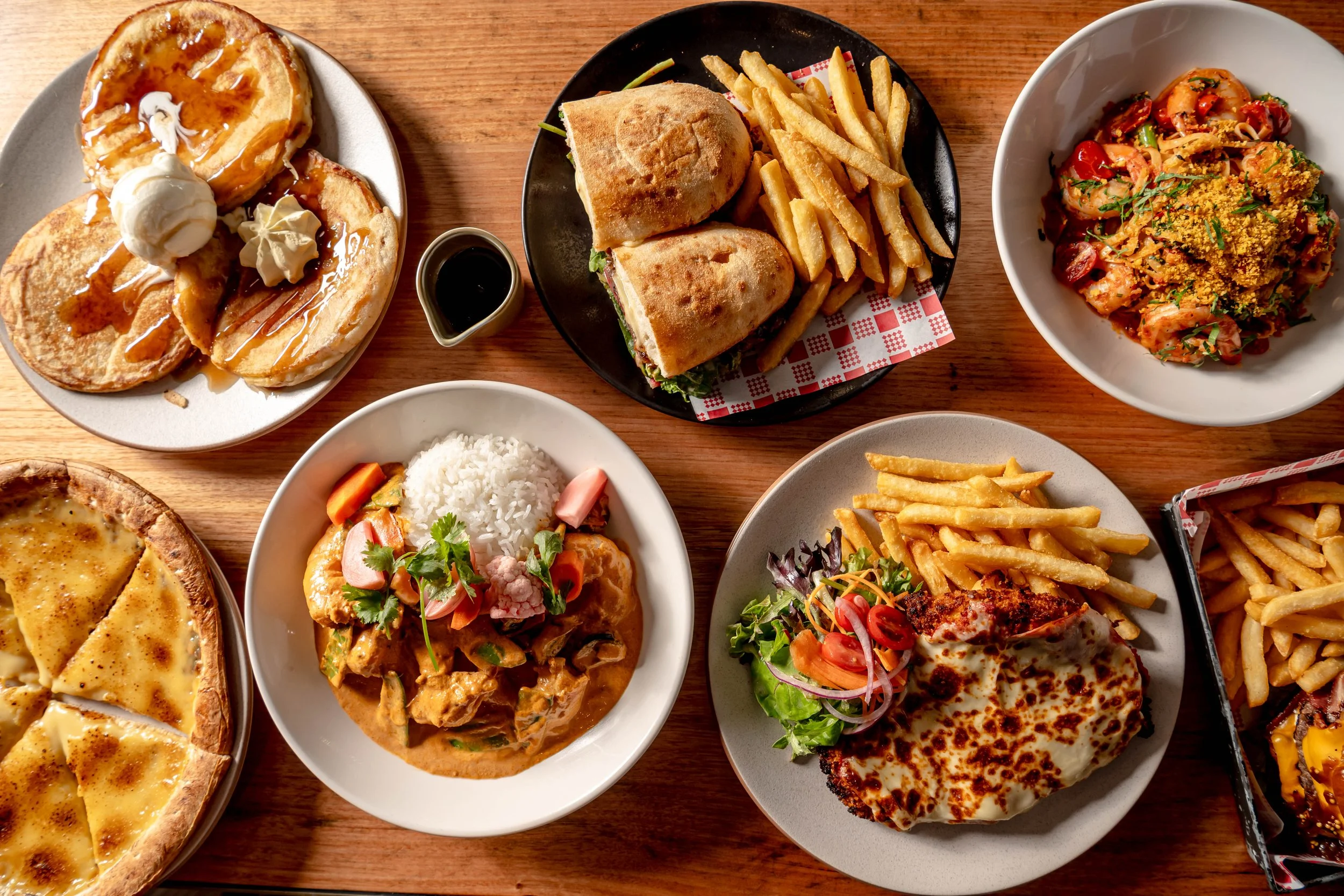 Assorted dishes of American comfort food on a wooden table including pancakes with syrup and ice cream, a chicken sandwich with fries, a bowl of shrimp pasta, a plate of cheese pizza, a plate with curry and rice, a chicken parmesan with salad and fries, and a burger with fries.